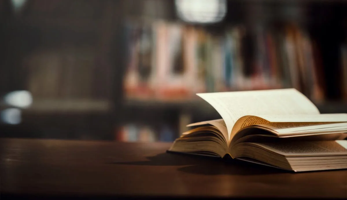 An open book on a wooden table in a dimly lit library or study, with bookshelves in the background.