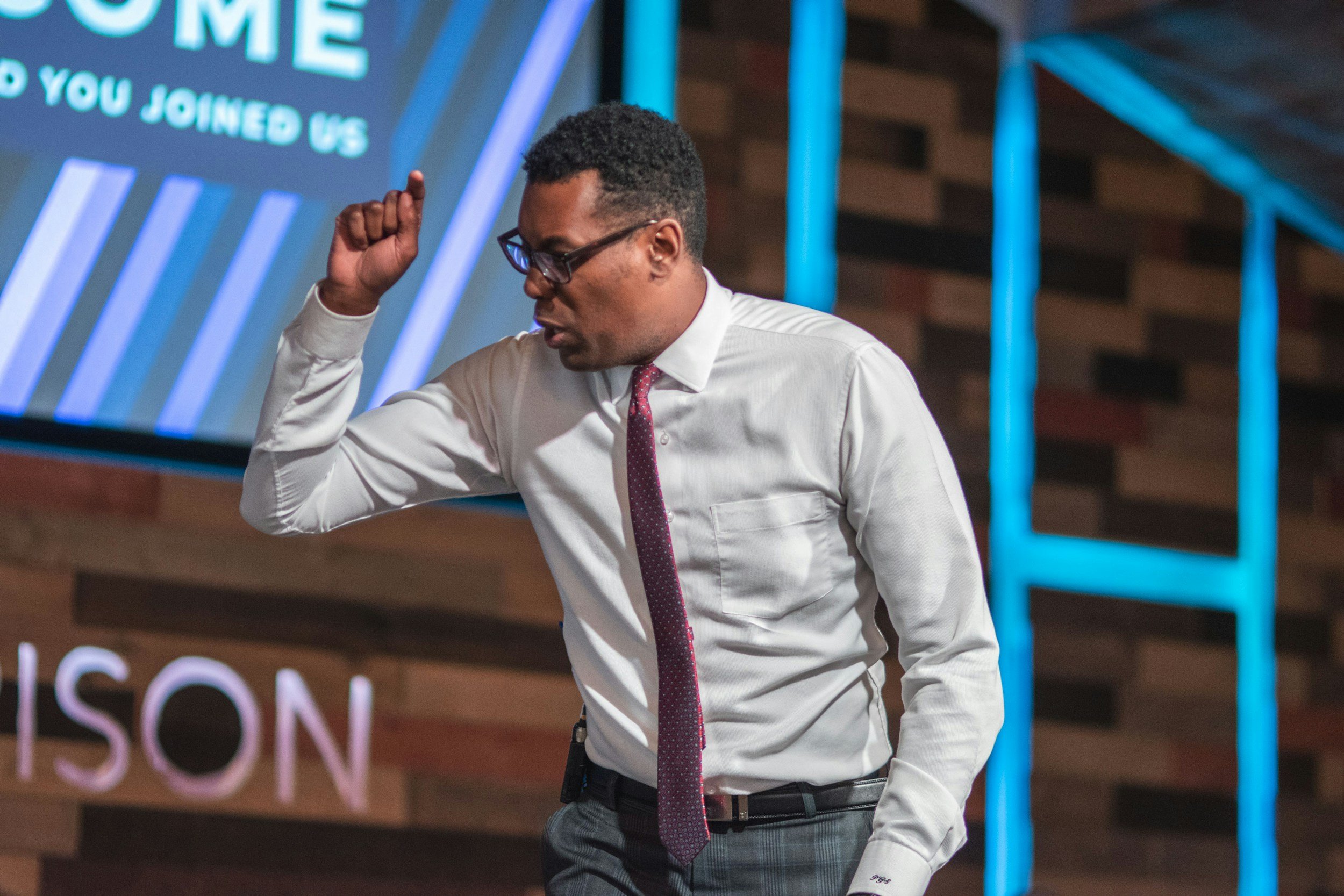 A man wearing glasses, a white dress shirt, and a pink tie is standing with his right hand raised to his head, possibly in a gesture of concentration or emphasis. The background includes a large signboard with partially visible text and blue vertical supports.