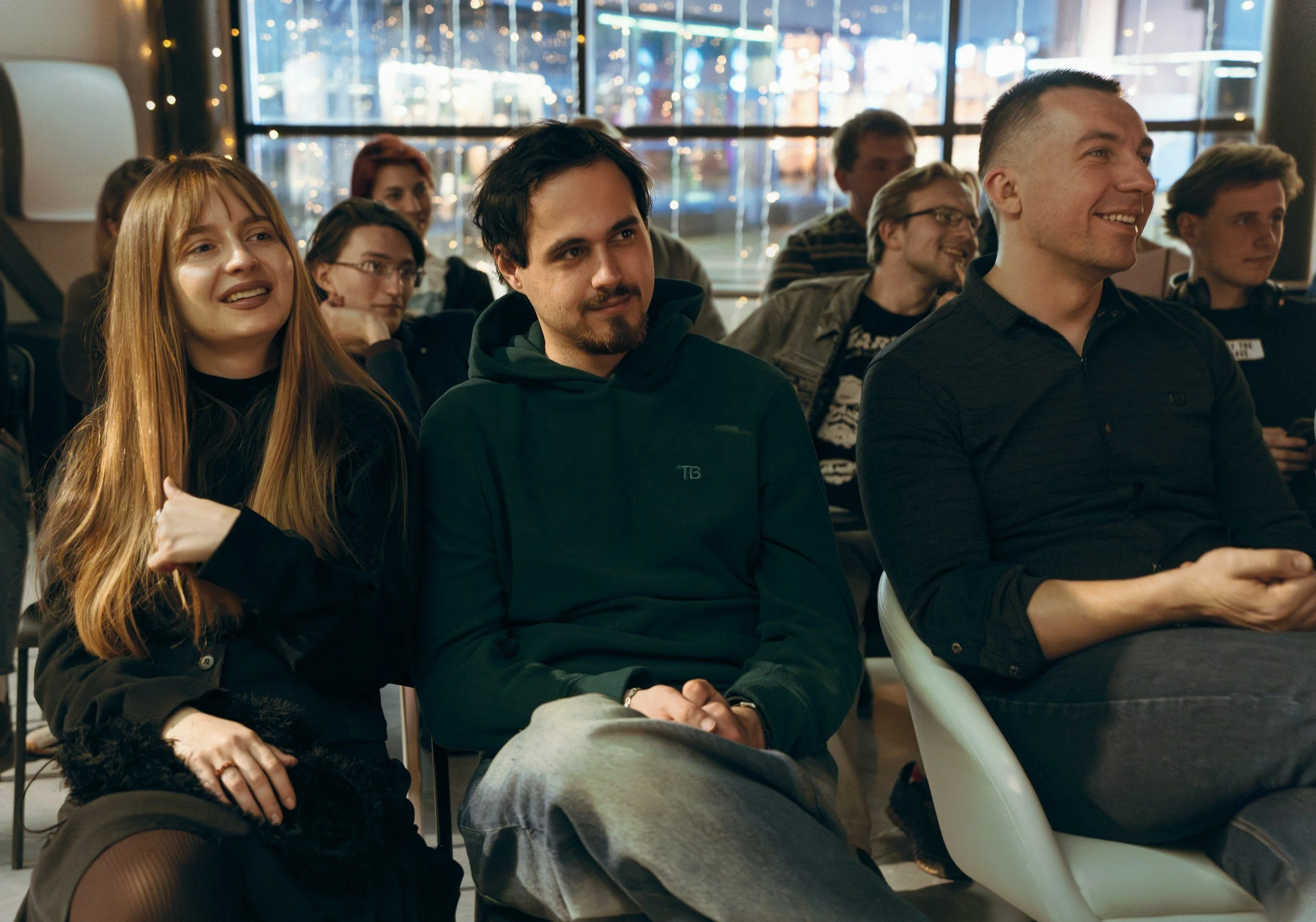 A group of young adults seated in a modern indoor space with large windows at night, some smiling and engaged, dressed casually.
