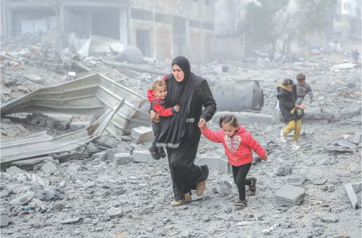A woman in a black headscarf and dress walking through a war-torn area with three children, holding a child's hand. The landscape around them is filled with debris and destroyed buildings.