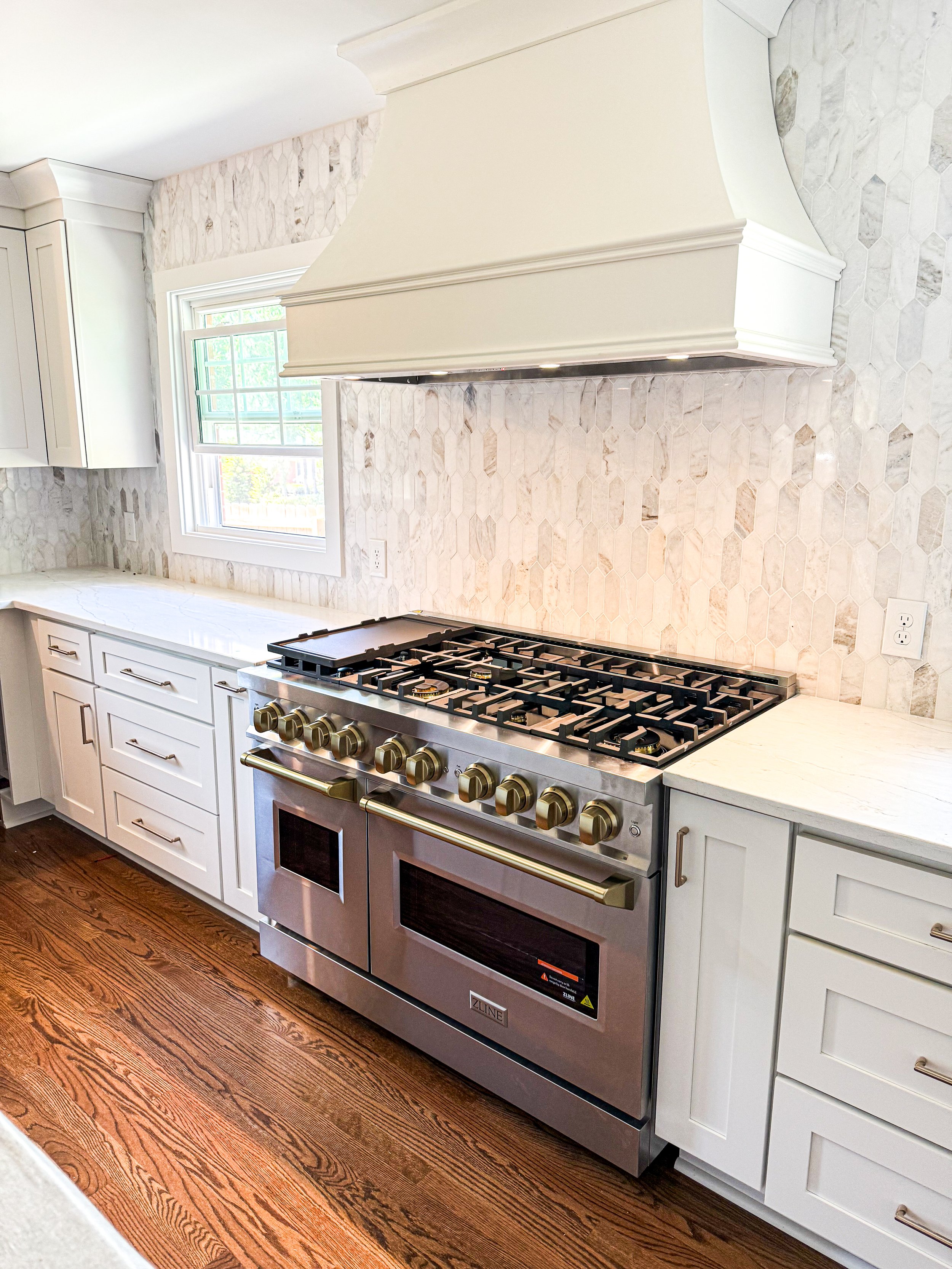 Kitchen with a modern stainless steel ZLINE gas oven, white cabinetry, a natural stone tile backsplash, and a window above the countertop.