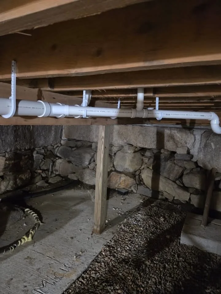 Underneath a wooden floor with plumbing pipes running along the joists, supported by vertical wooden posts, with a stone wall in the background and gravel ground below.