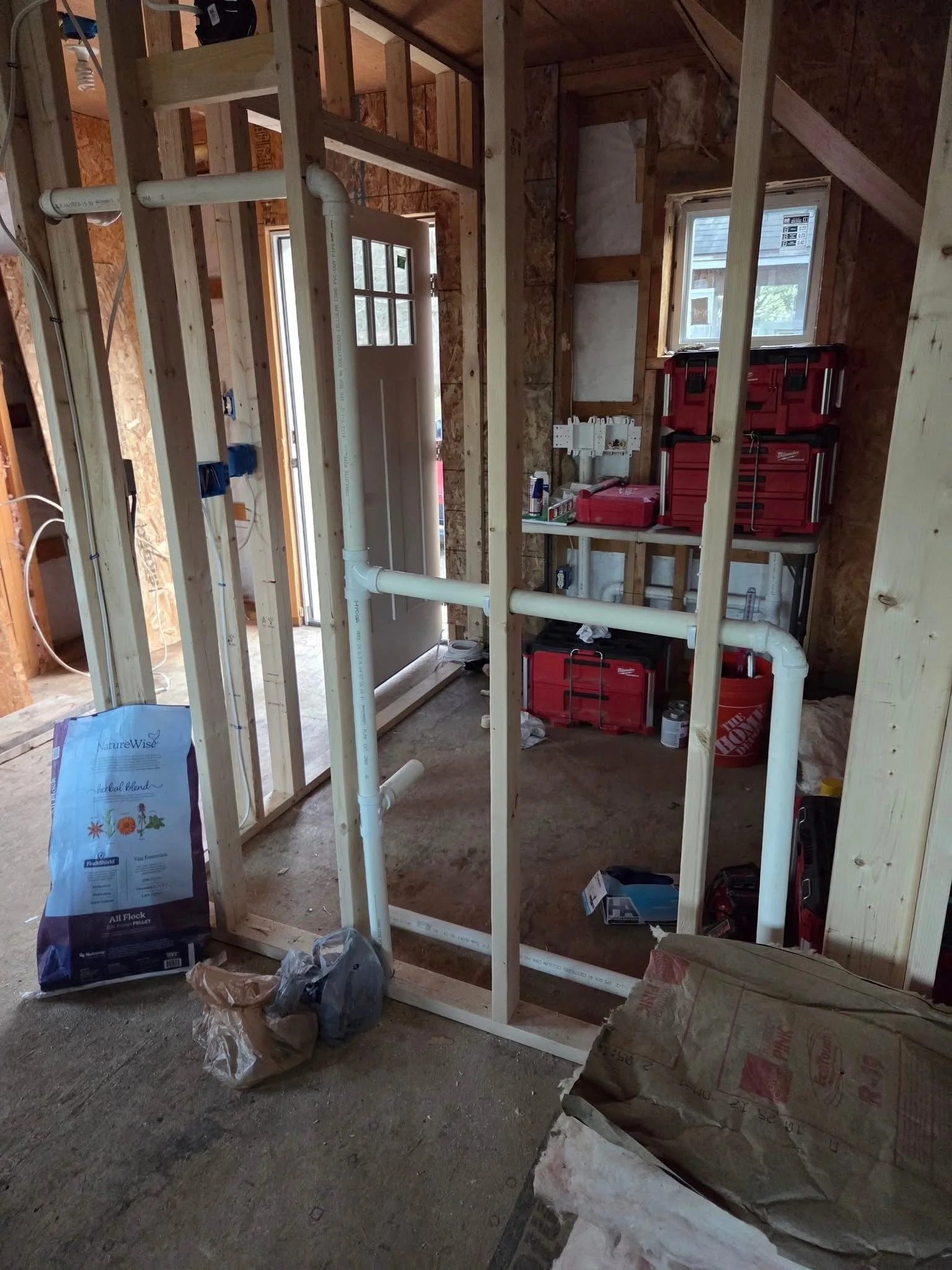 Interior of a house under construction with exposed wooden framing, PVC piping, a window, tools, and construction materials.