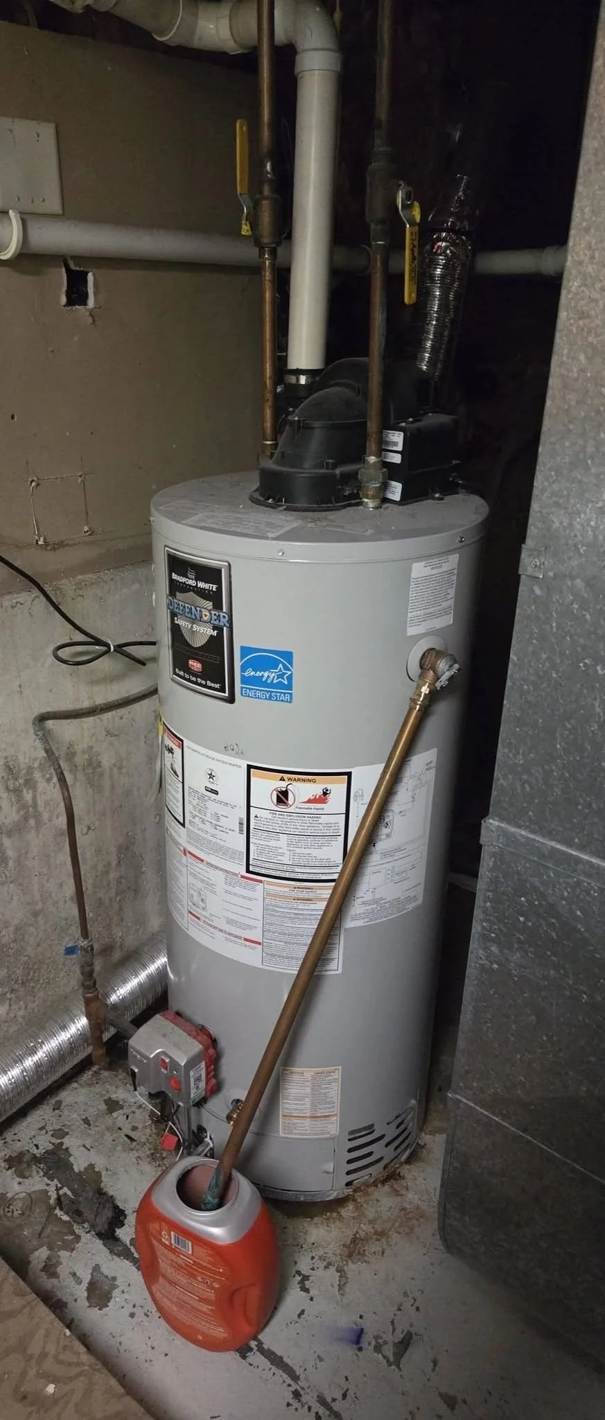 A water heater installed in a basement with pipes, vents, and an extension cord nearby, along with a red container. The water heater has energy efficiency labels and safety warnings.