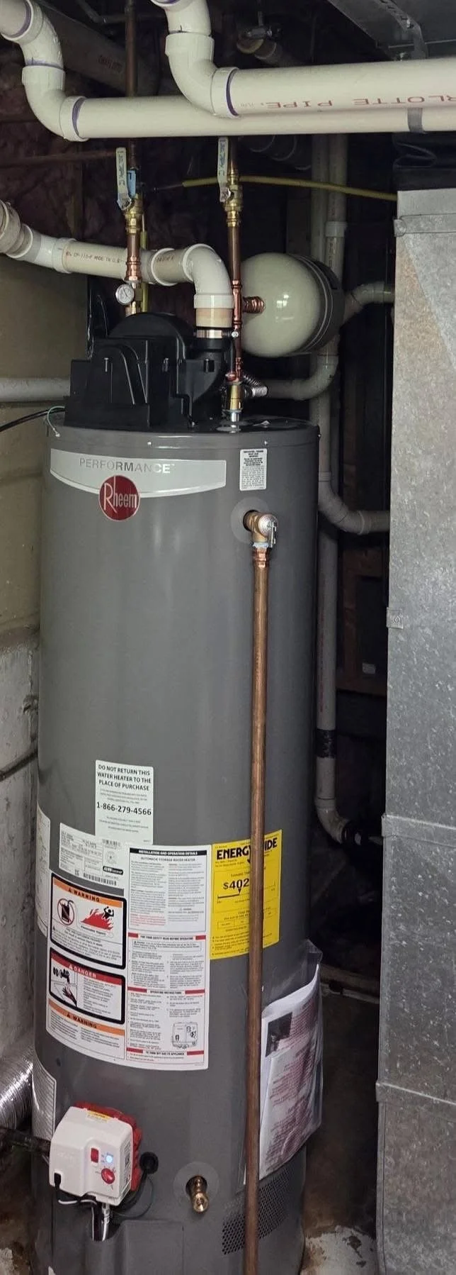 A Rheen Performance water heater system with copper and PVC pipes connected to it, situated in a utility room. Pipes are labeled with blue and red, indicating cold and hot water lines. The heater has warning and energy guide labels.