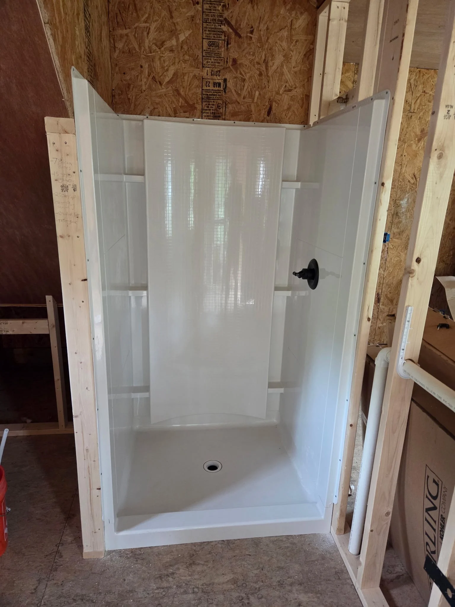 Image of a partially constructed bathroom shower with white tile walls, a black showerhead, and wooden framing, set in an unfinished room with exposed plywood and construction materials.