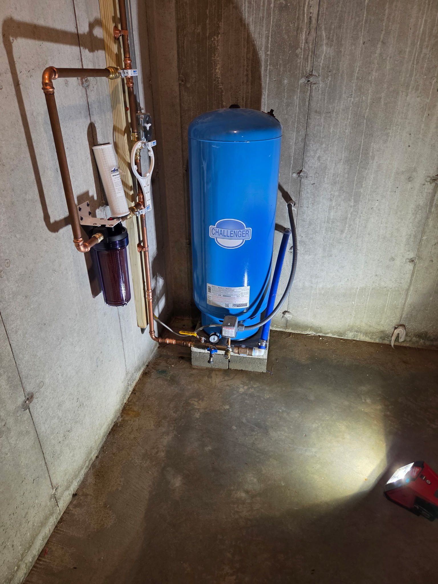 A blue Challenger water pressure tank connected to copper pipes, a filter, and a pressure gauge in a concrete basement.