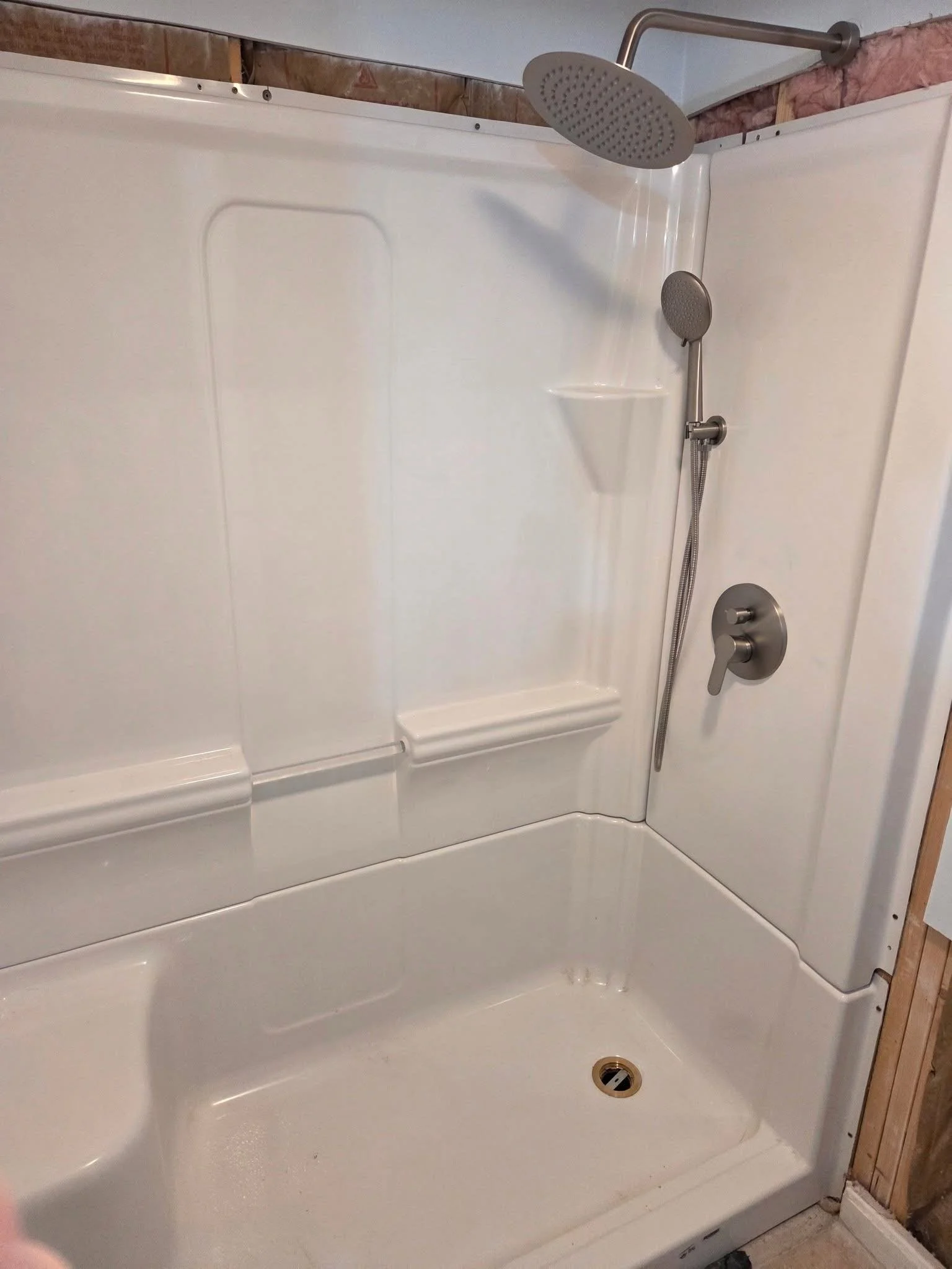 A white shower with a built-in seat, a handheld shower head, and a rain shower head mounted on the ceiling, with exposed wall studs above.