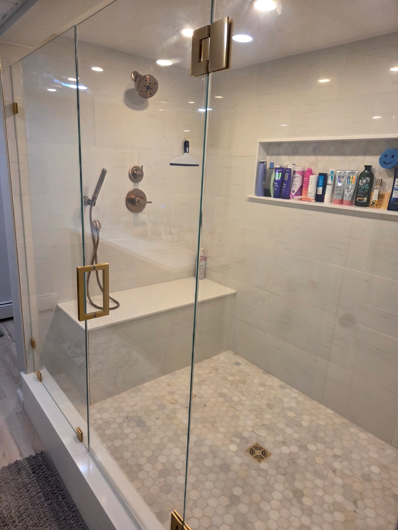A modern walk-in shower with a glass door and gold handles, featuring a rainfall showerhead and a handheld shower. The shower has a built-in shelf with toiletries and a tiled wall with small hexagonal tiles. The shower floor has hexagonal tiles and a