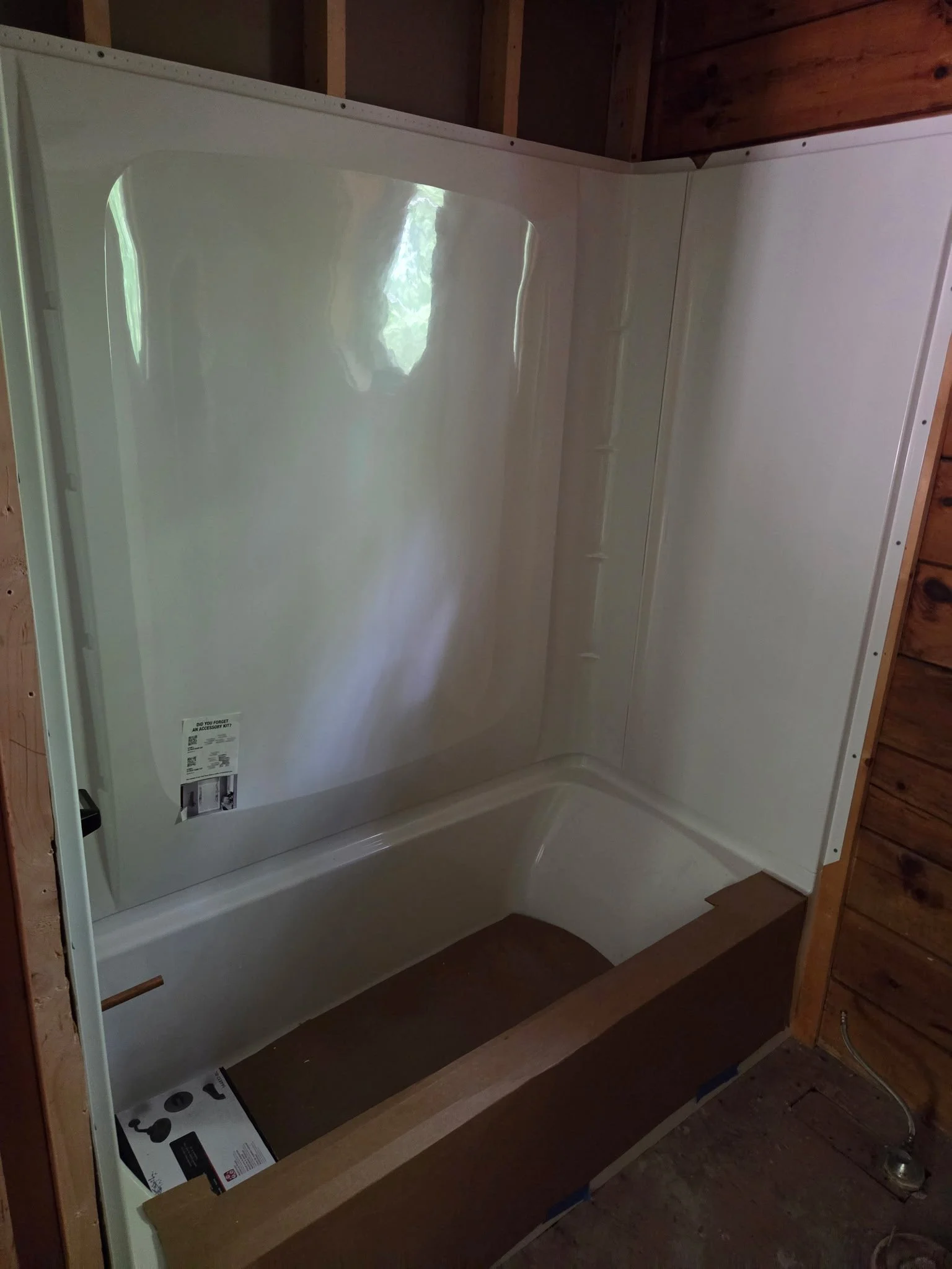 A white shower stall with a curved ceiling, installed in a wooden-framed bathroom. The stall has a clear plastic footer on the floor and a sticker on the side.