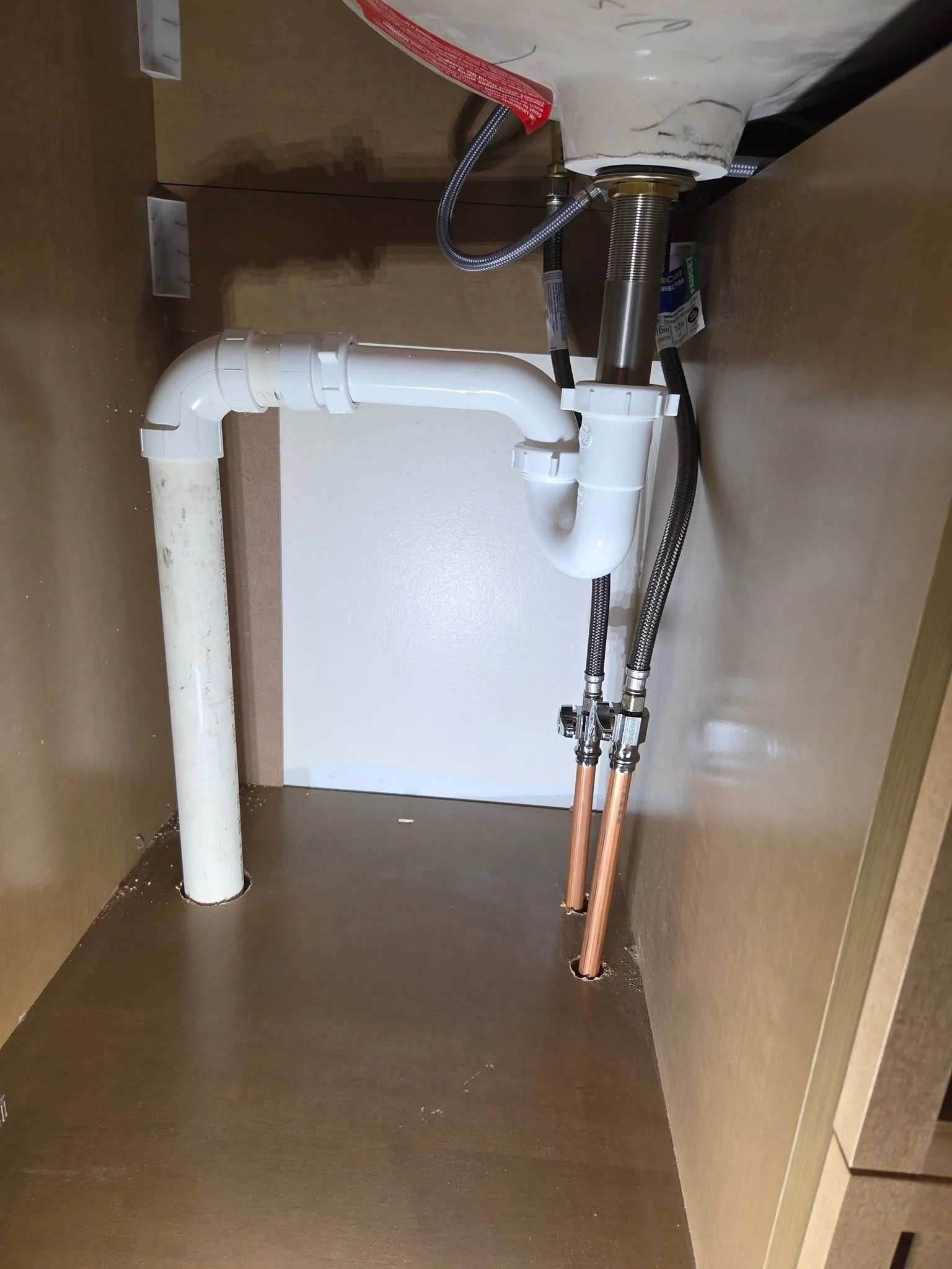 Underside view of a kitchen sink area showing plumbing pipes, drain connections, and flexible water supply lines, with a brown cabinet and a wall in the background.