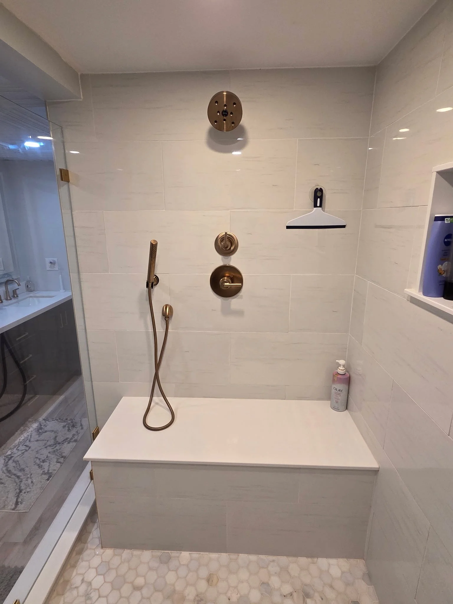 Shower area with beige tiled wall, brass fixtures, a hand-held showerhead, a control handle, a soap shelf, a wall-mounted squeegee, and a bottle of Olay body wash.