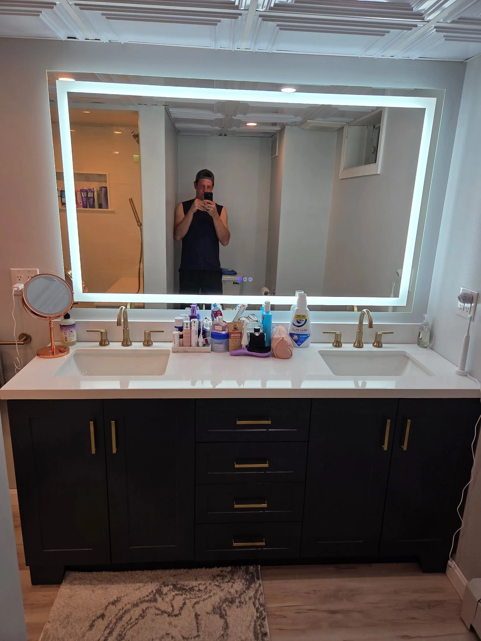 A bathroom vanity with a double sink, dark cabinet, gold handles, and cluttered countertop with toiletries. A large illuminated mirror with a white frame is above the vanity, reflecting a man taking a photo with his phone. The bathroom has a beige an