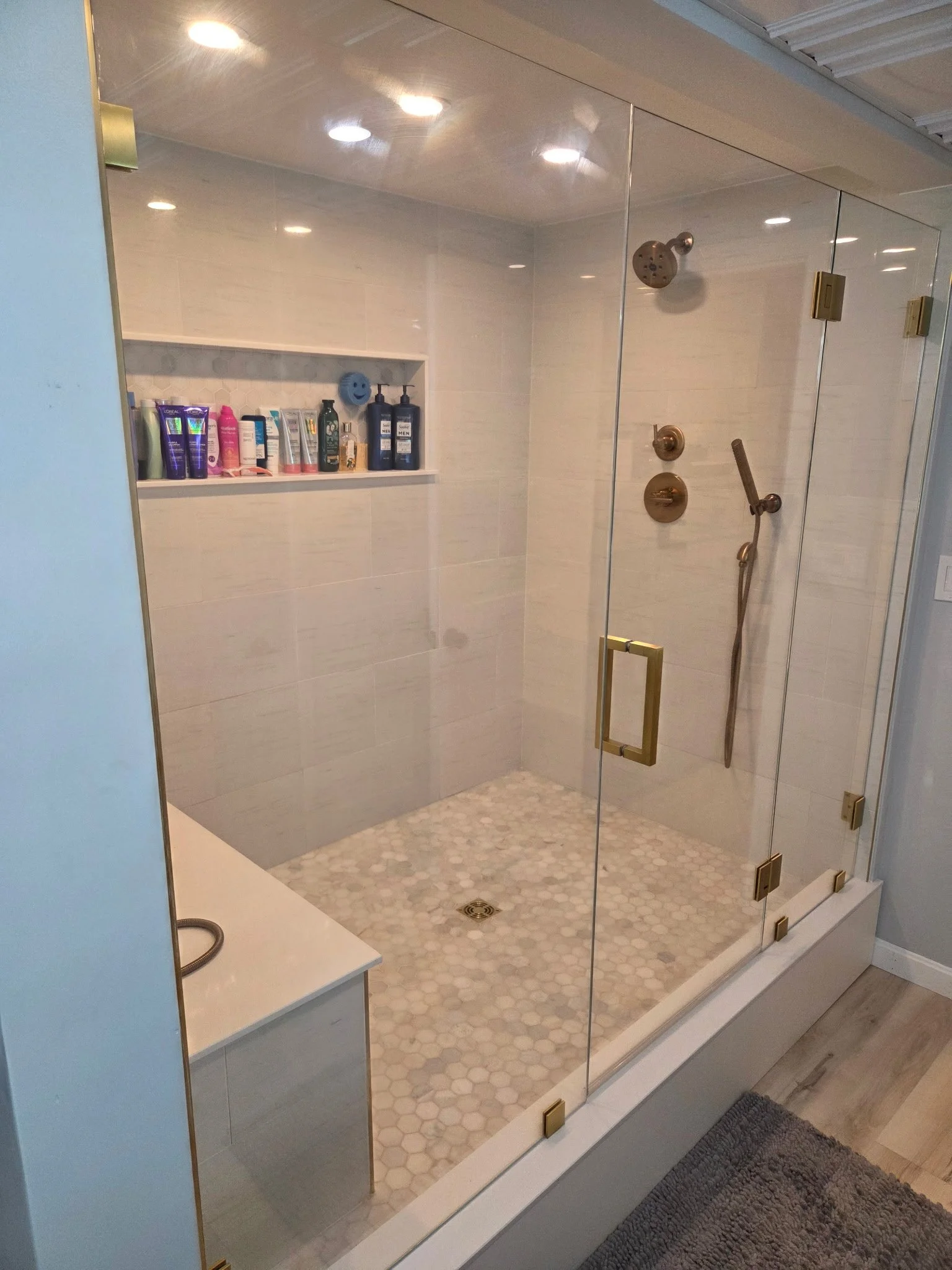 Modern walk-in shower with glass enclosure, brass fixtures, and beige tile walls and hexagonal tile flooring. Contains a built-in shelf with toiletries and a handheld showerhead.
