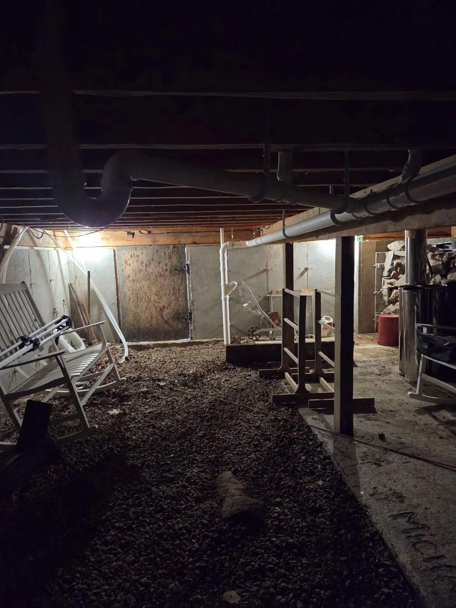 Underground construction site with exposed pipes, white chairs, and construction materials, illuminated by a single light, with a gravel floor.