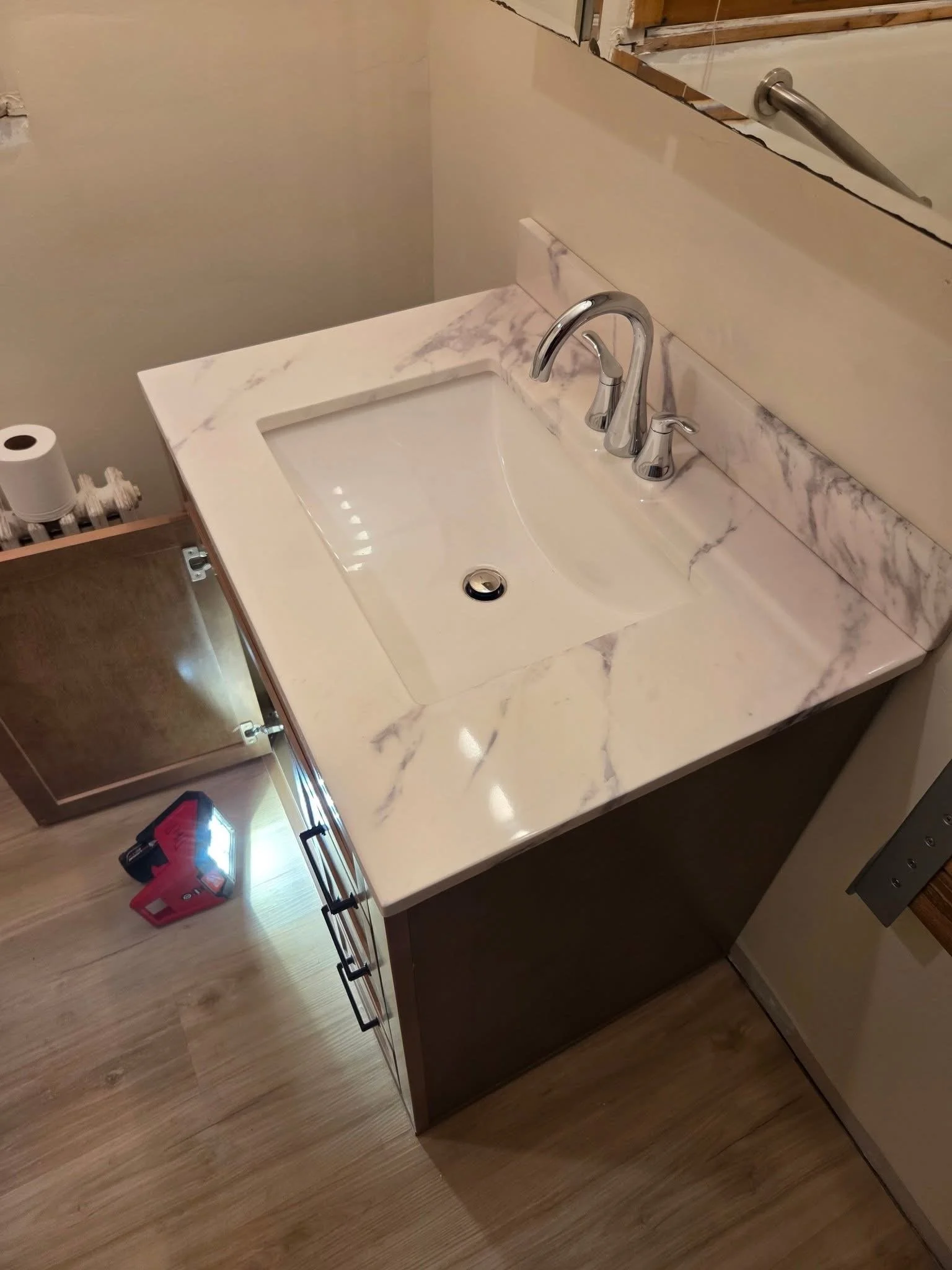 A bathroom vanity with a marble countertop, a sink, and a chrome faucet. There is a wall mirror above the sink. A cabinet with a mirror door is partially visible on the left. The bathroom floor is wooden.