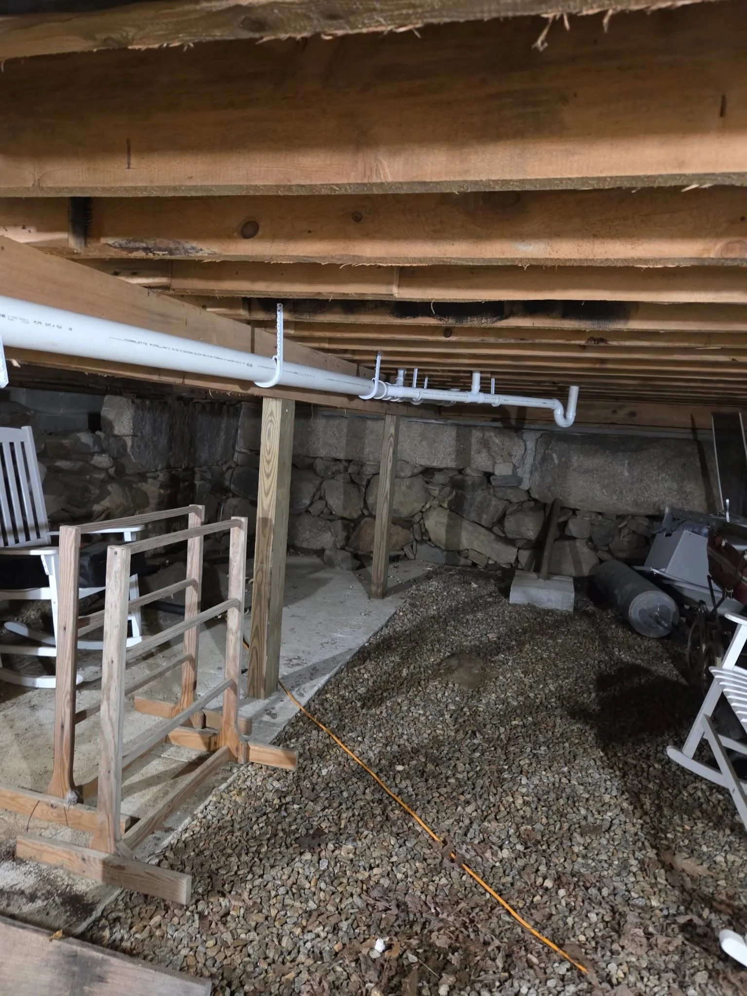 Photo of basement with wooden beams on ceiling, PVC pipes installed along the ceiling, and a gravel floor. There are some chairs and construction materials visible.