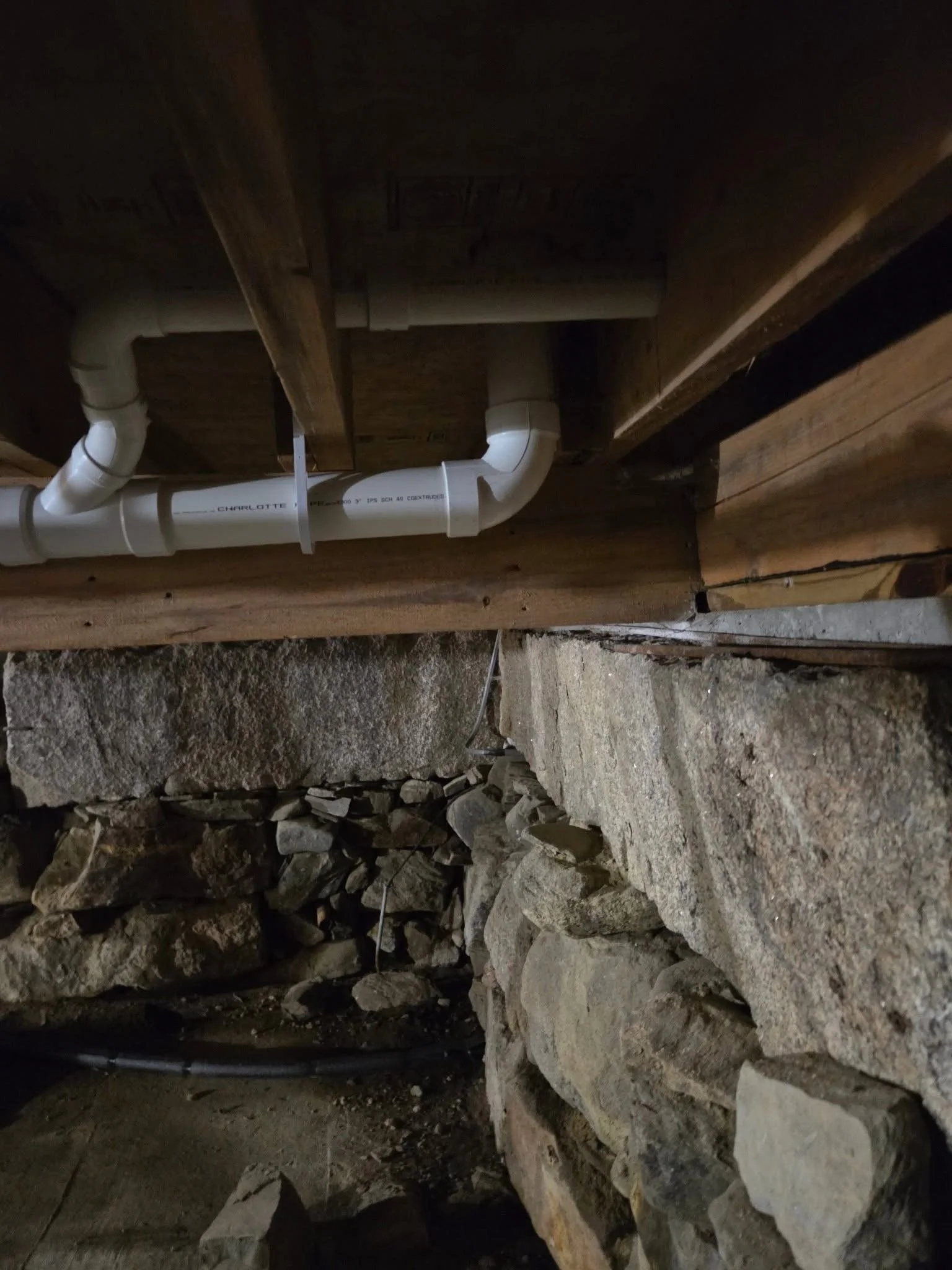 Underneath a wooden structure with visible pipes and rough stone ground, construction or plumbing work is underway.