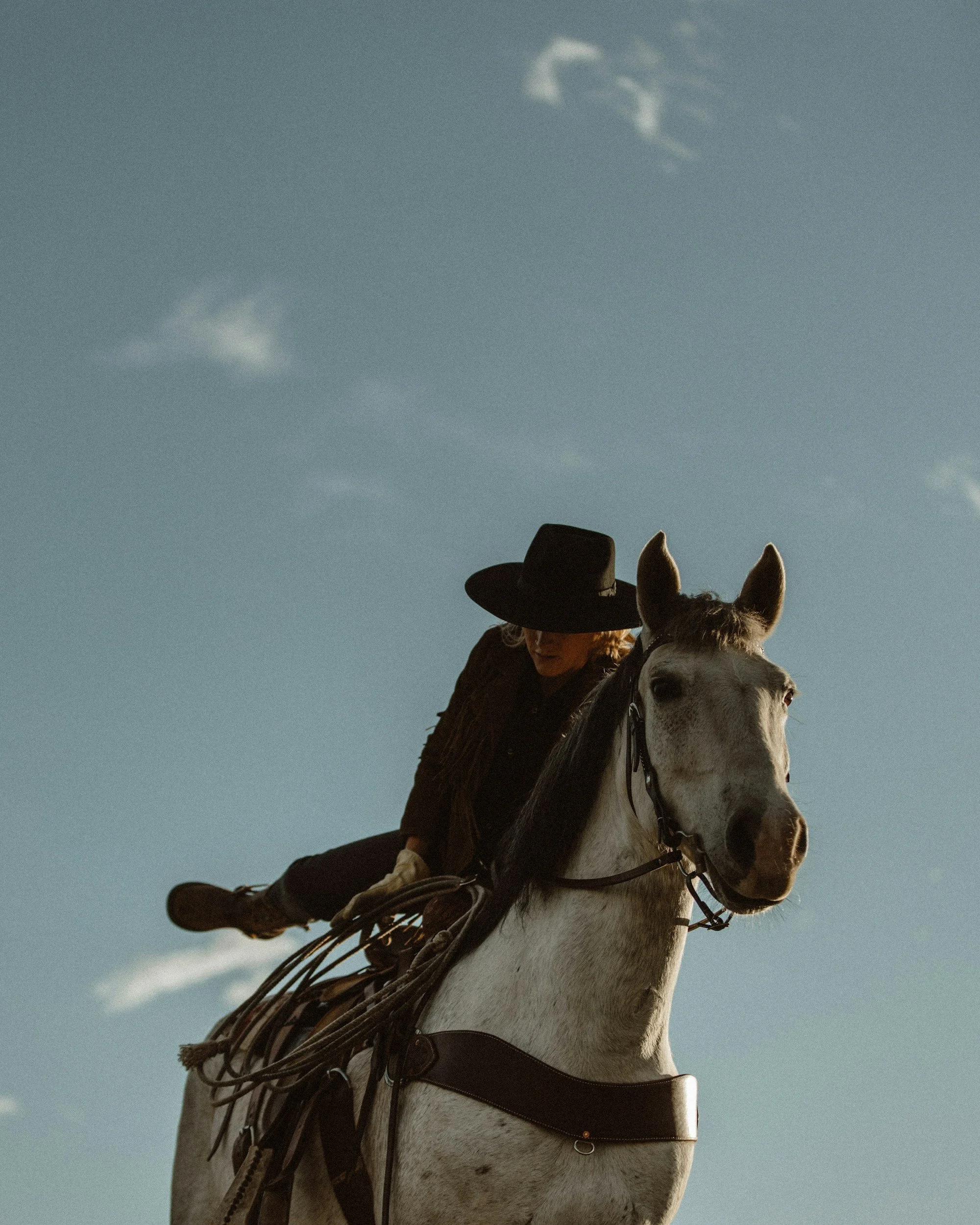A person wearing a wide-brimmed hat riding a white horse with a dark saddle and bridle, under a blue sky with a few clouds.