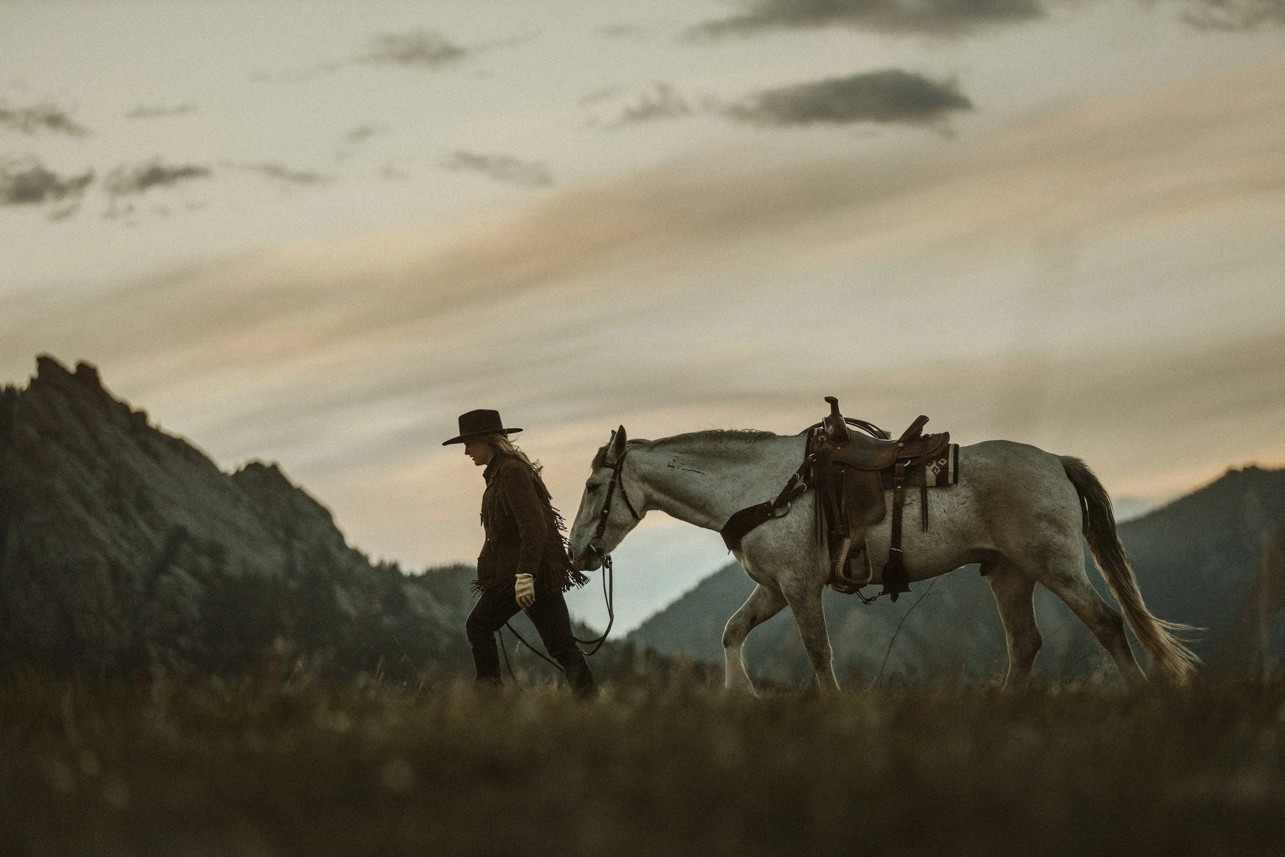 A woman walking a white horse in a mountainous landscape during dusk, wearing a hat and Western-style attire.