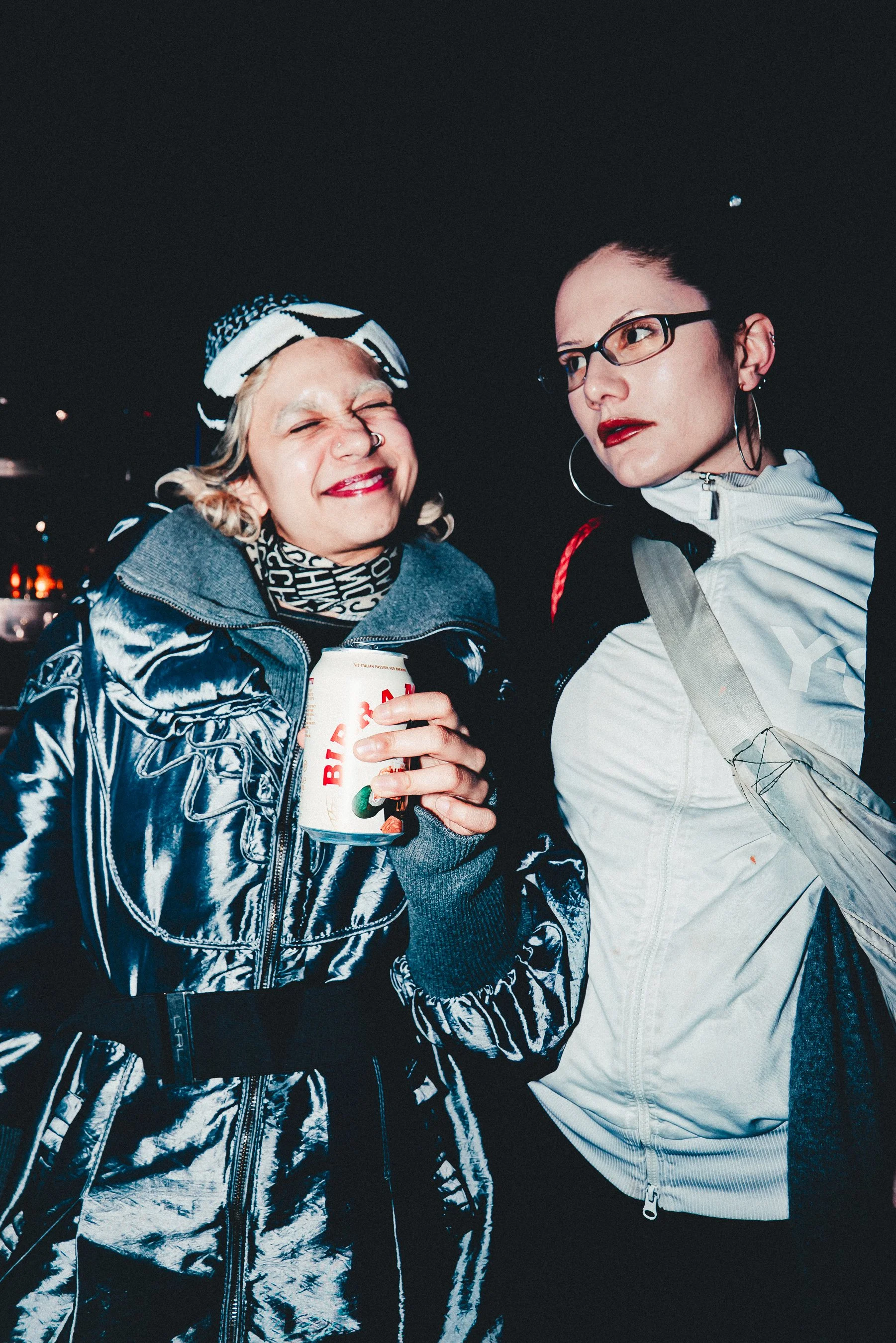 Two women at night, one with platinum blonde hair wearing a metallic jacket and a patterned headscarf, holding a can of Bud Light beer and smiling with eyes closed. The other woman has dark hair in a bun, wearing glasses, hoop earrings, a white jacke