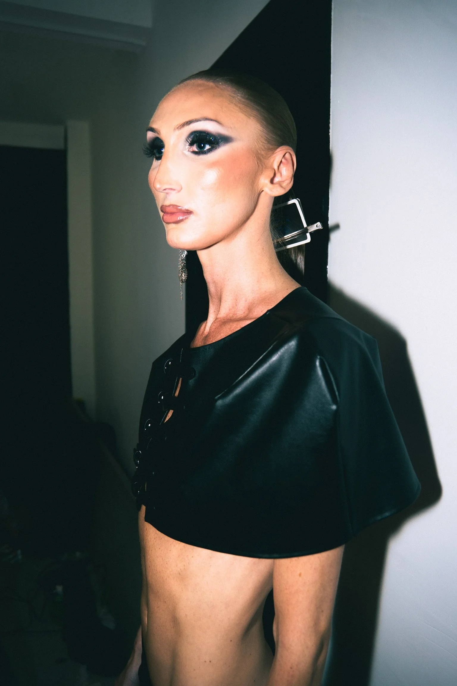 A person with a side profile in a black leather top, large hoop earrings, and dramatic makeup, standing against a plain wall.