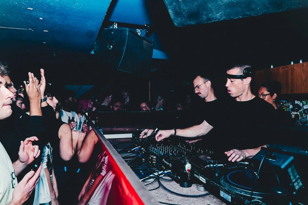 Two DJs perform at a club with a crowd watching, DJ equipment, and speakers in the background.