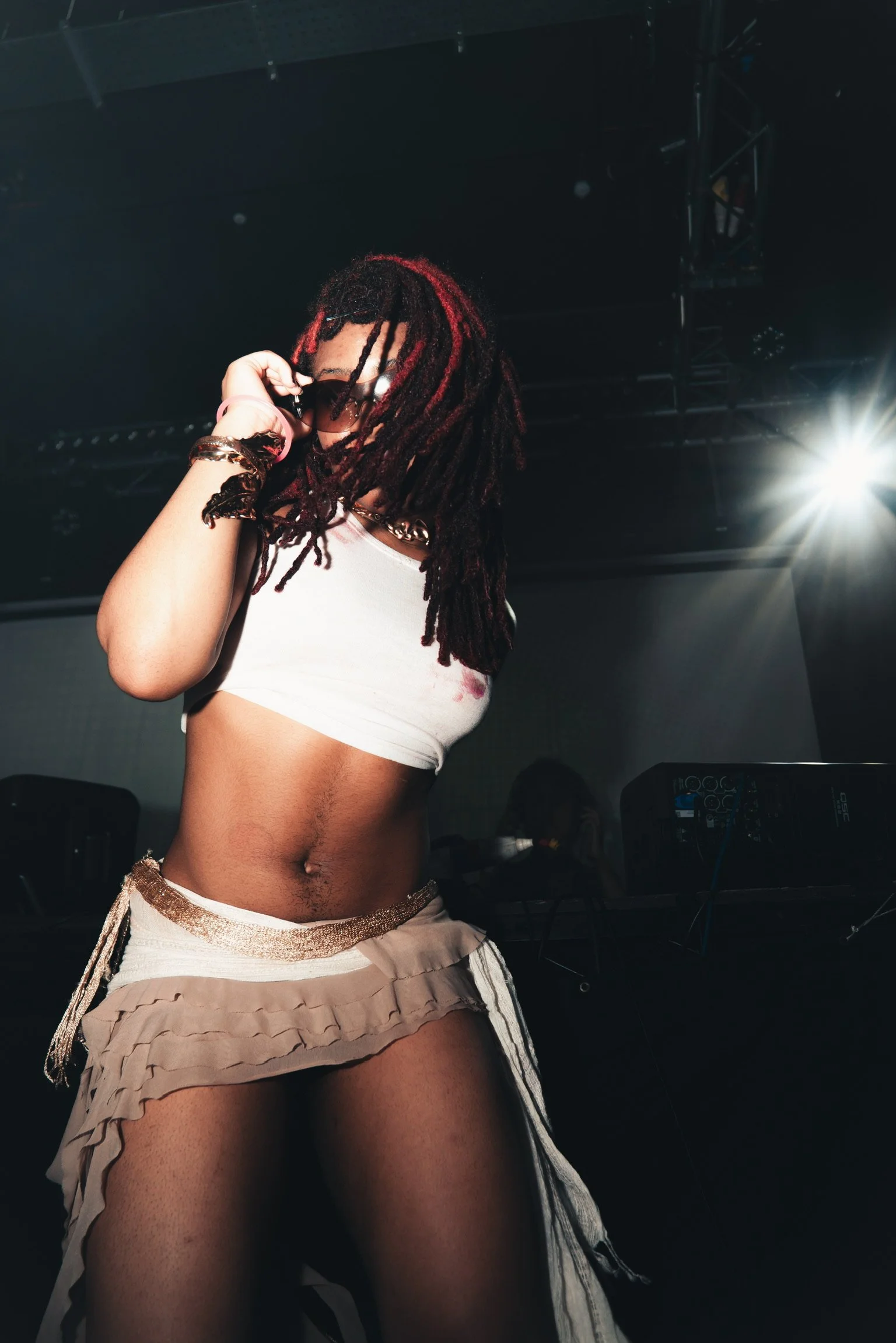 A woman with dreadlocks wearing sunglasses and a white crop top, standing on a stage with a DJ booth and lighting equipment in the background.
