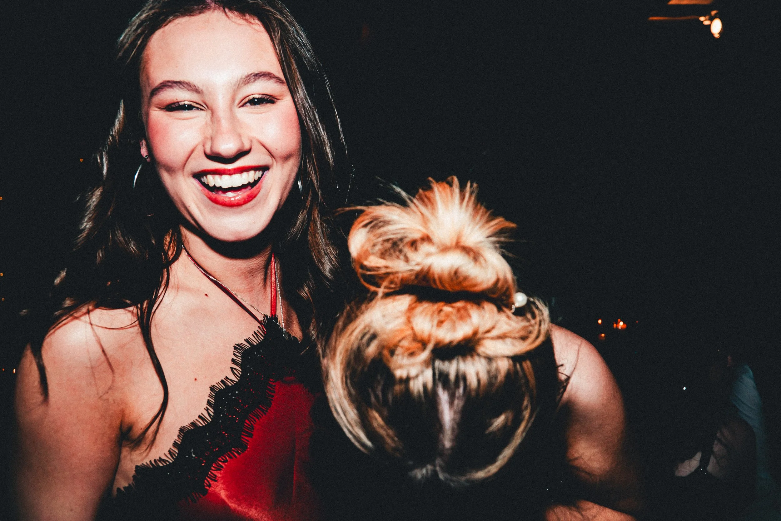 A woman with long brown hair, red lipstick, and earrings is smiling and holding a small dog with tan and black fur in front of a dark background with some lights.