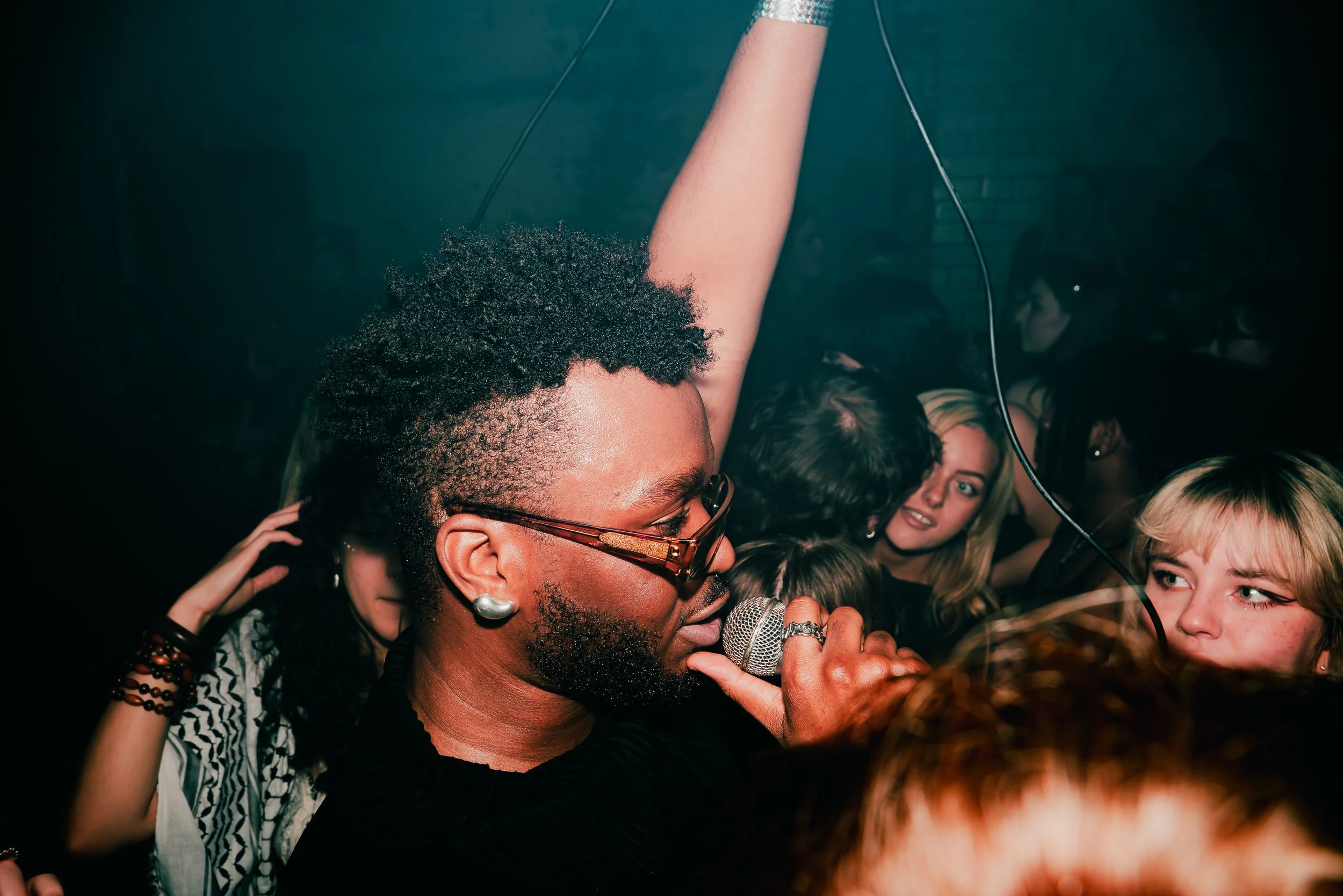 A singer with curly hair, glasses, and earrings performing with a microphone surrounded by a crowd at a lively event.