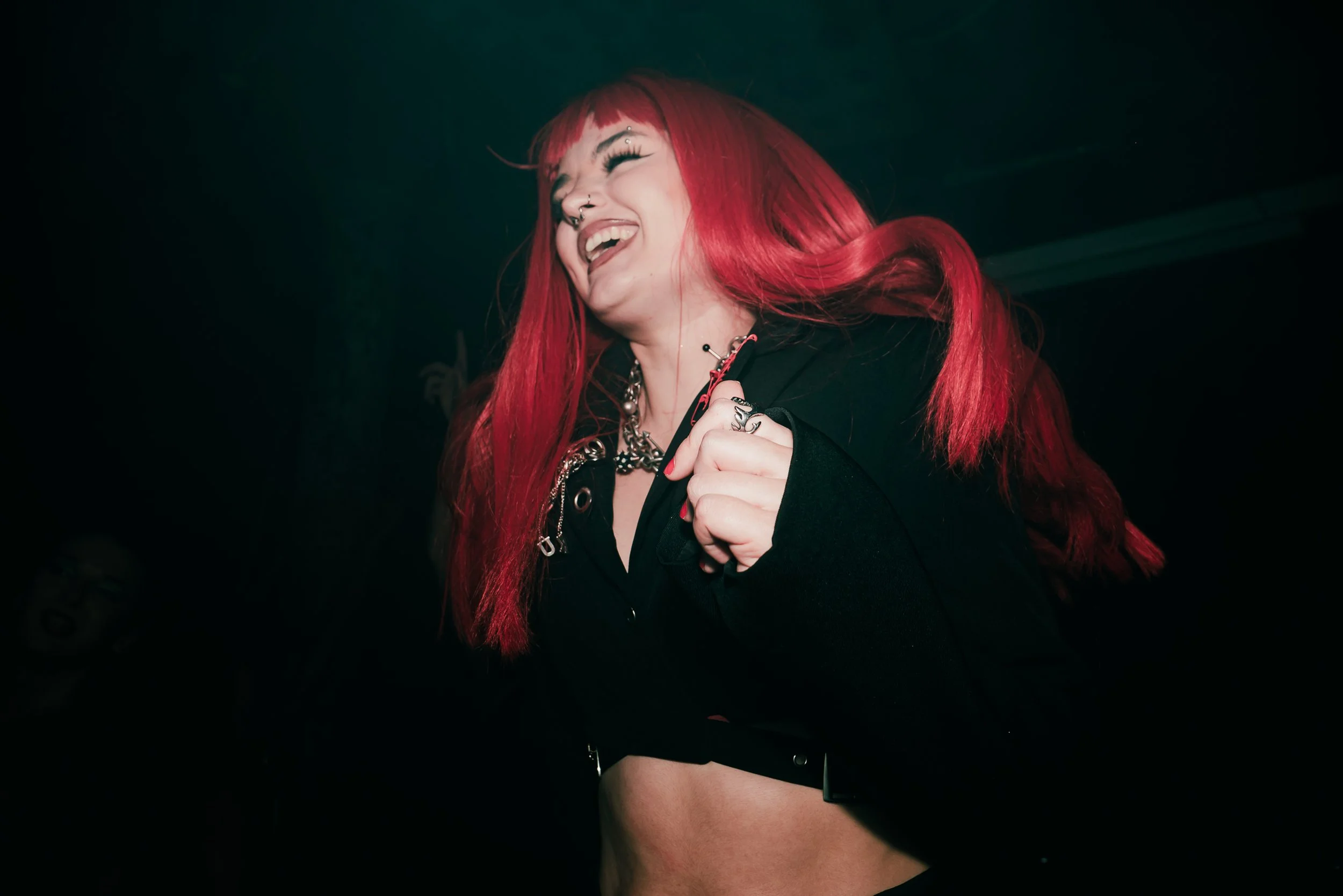 A woman with bright red hair laughing, wearing dark clothing and jewelry, in a dark setting.