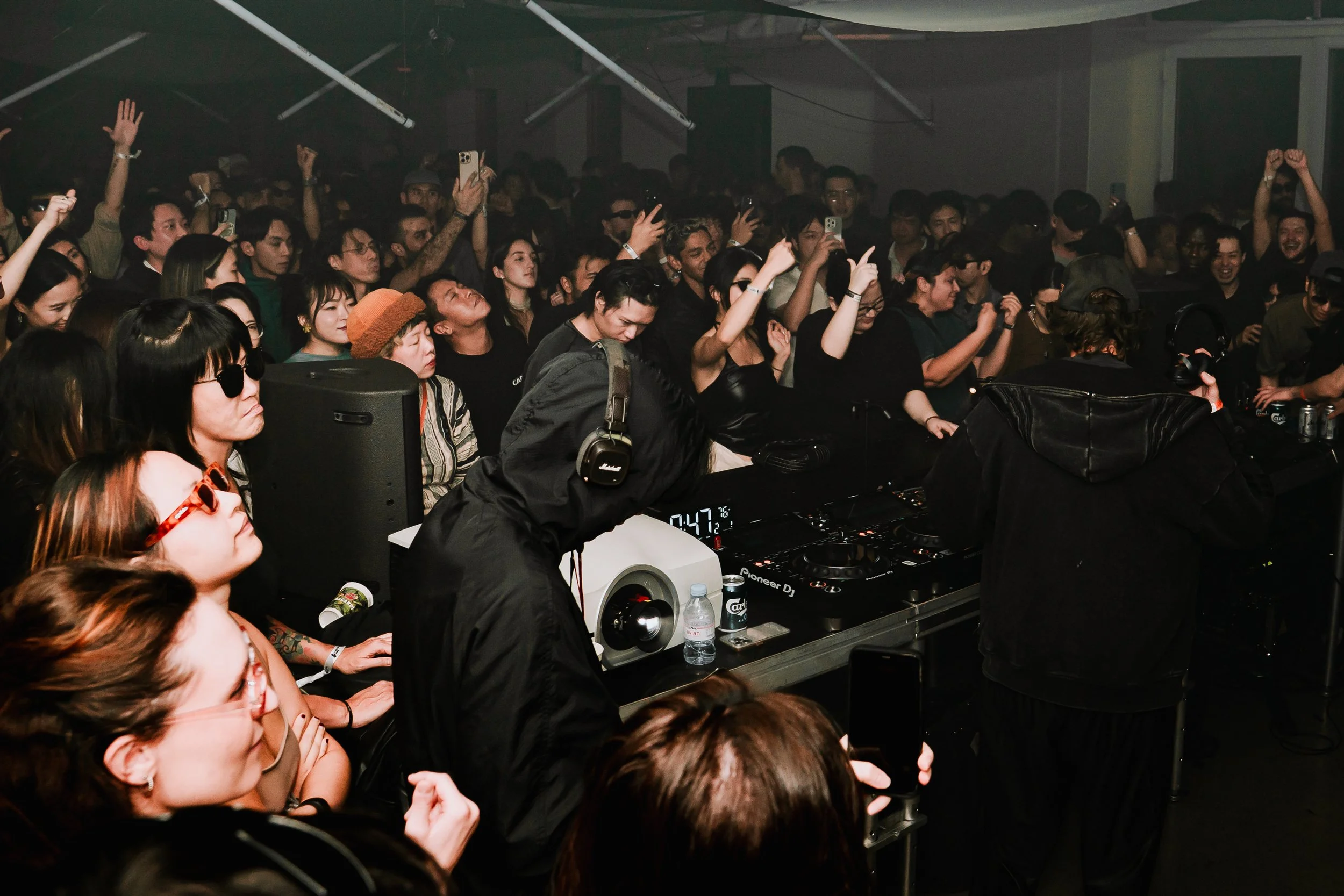 A crowded indoor concert or club scene with many people dancing and enjoying music. The crowd features diverse individuals, some with glasses, tattoos, and headphones, with DJ equipment visible in the foreground.