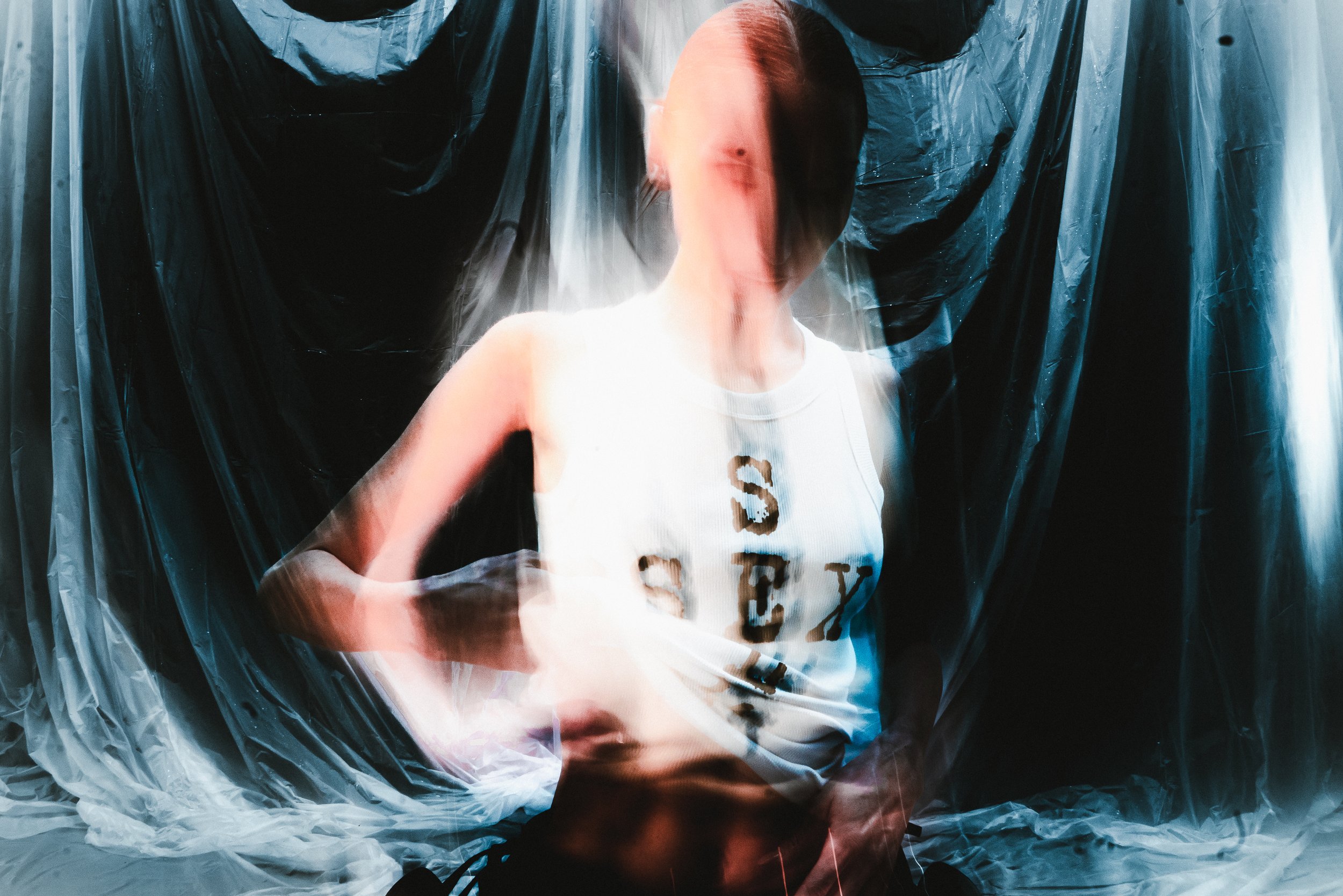 A person wearing a white sleeveless T-shirt with the letter 'S' on it, standing in front of black plastic and transparent plastic sheets.