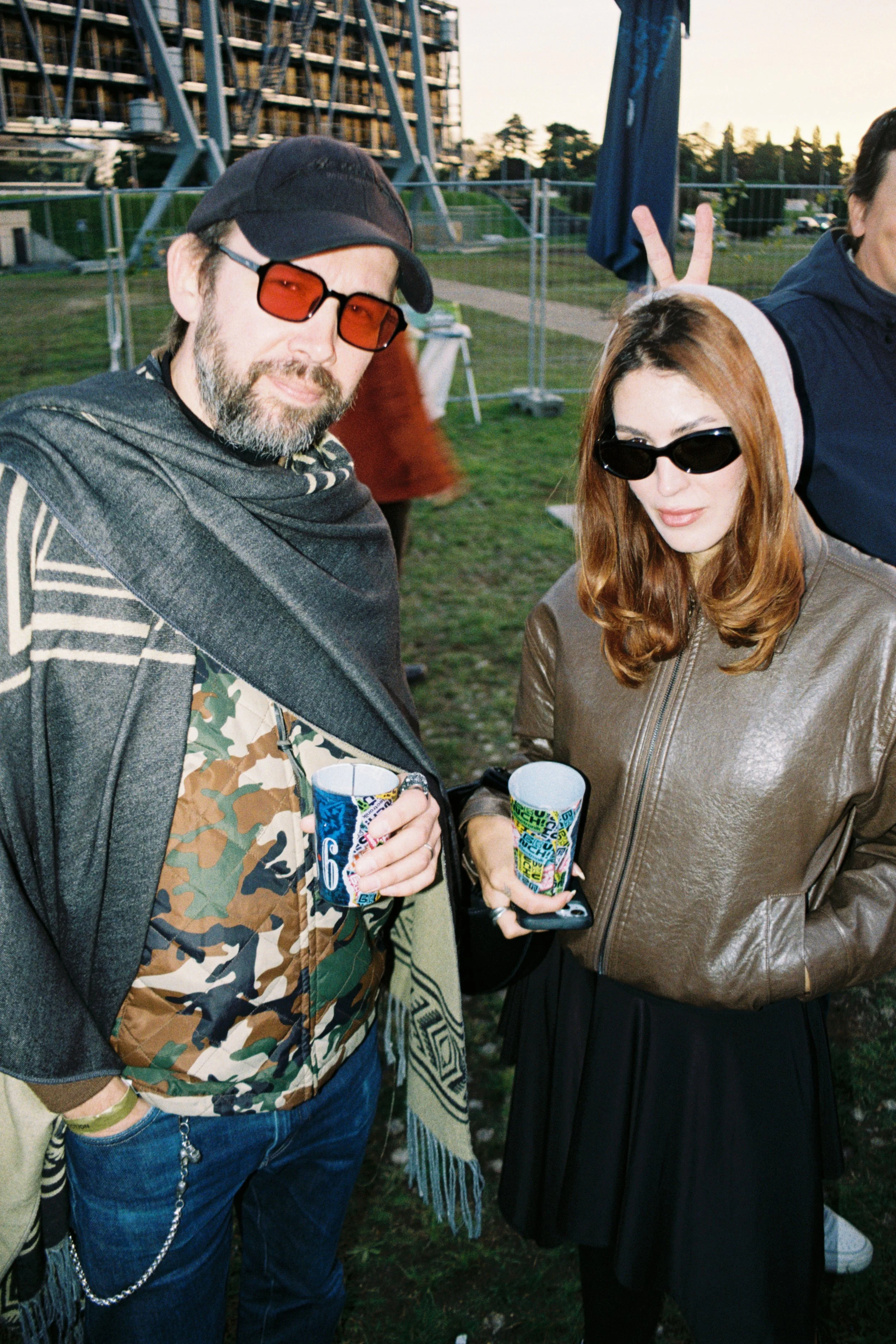 Two people holding colorful cups at an outdoor gathering during sunset, with a roller coaster structure and trees in the background.