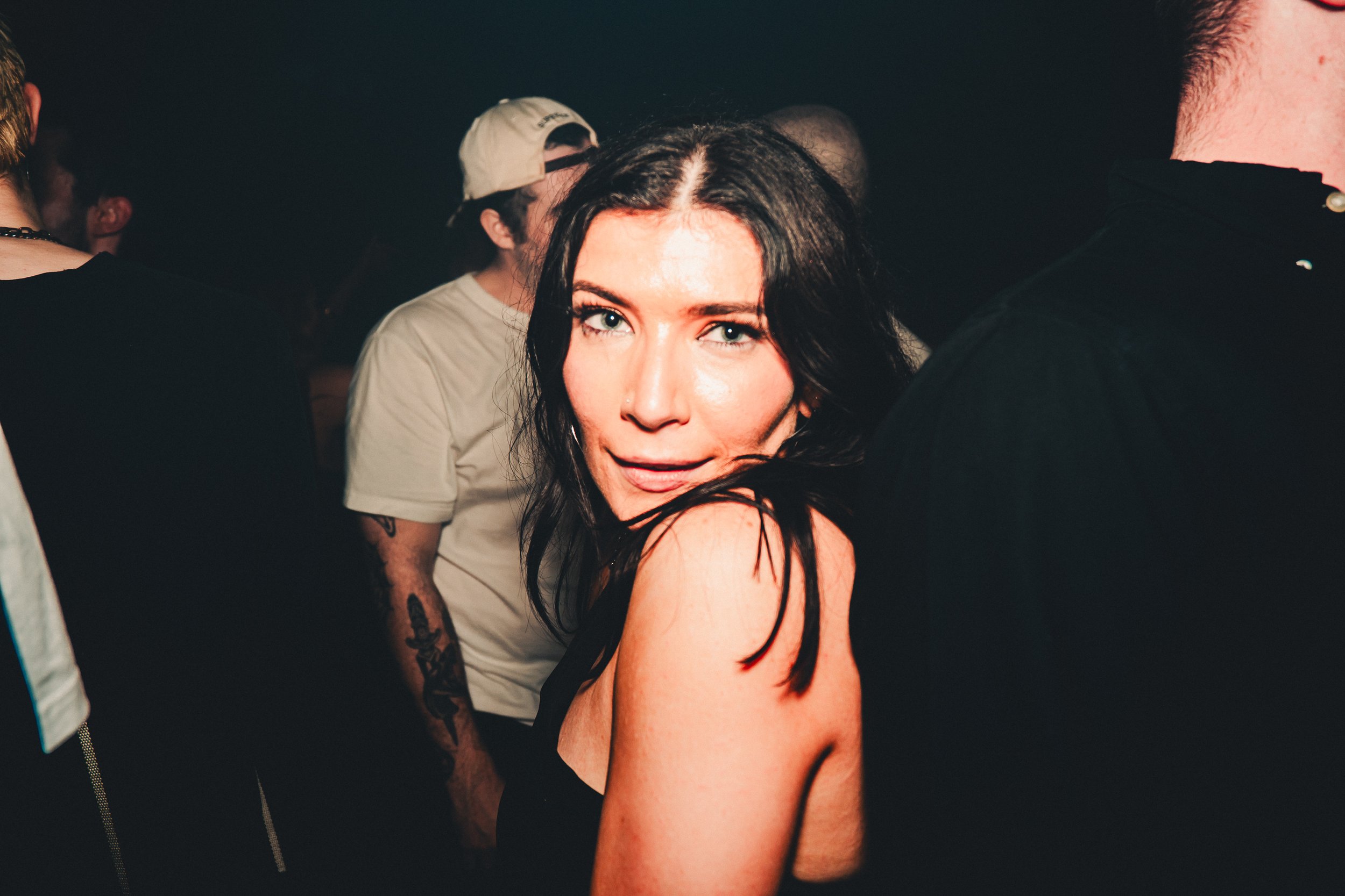 A woman with black hair and blue eyes looking at the camera, surrounded by people in a dimly lit setting, possibly a party or nightclub.