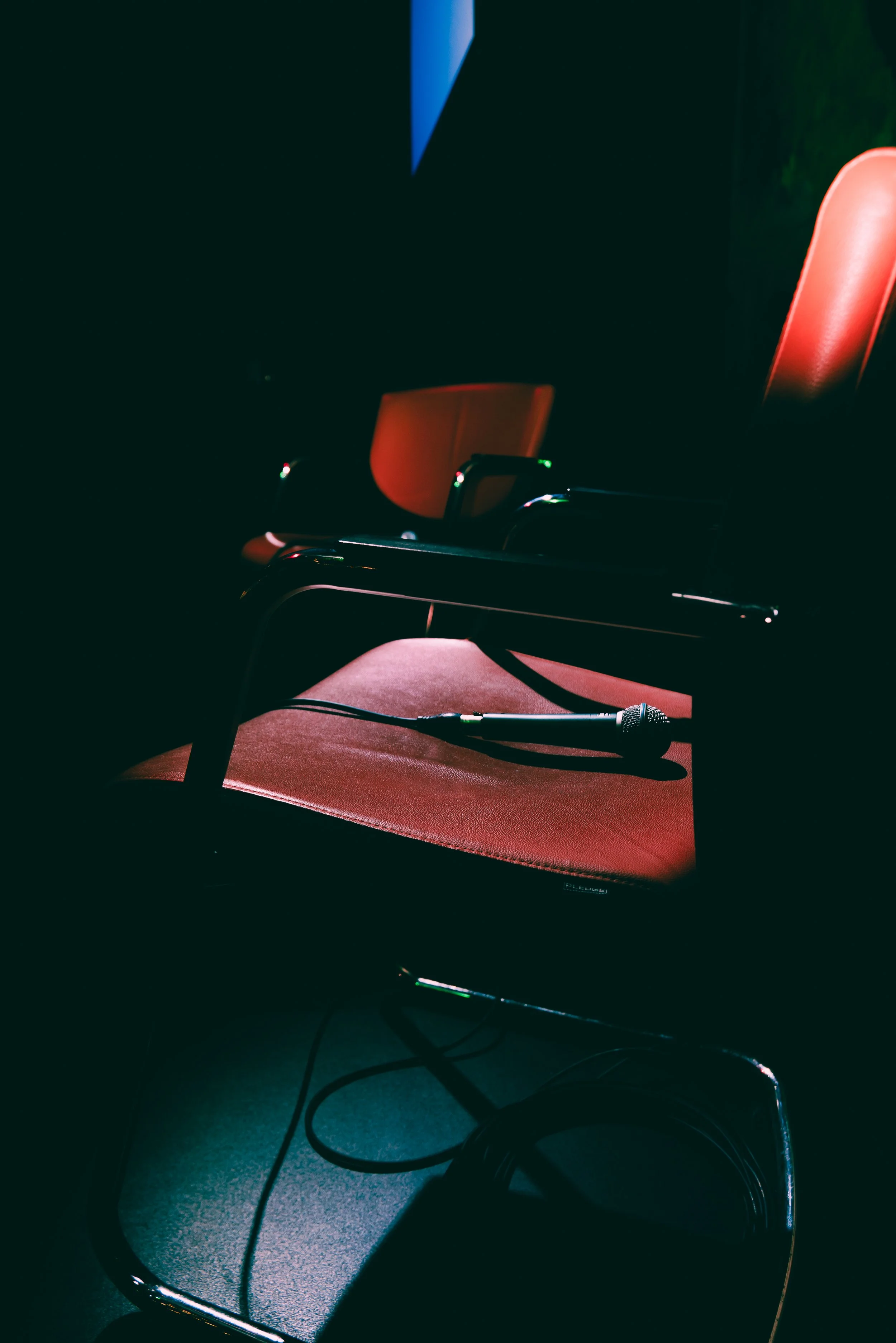 Dimly lit scene with a red chair, a microphone, and some cables on the floor, suggesting a music or speaking setup in a dark room.