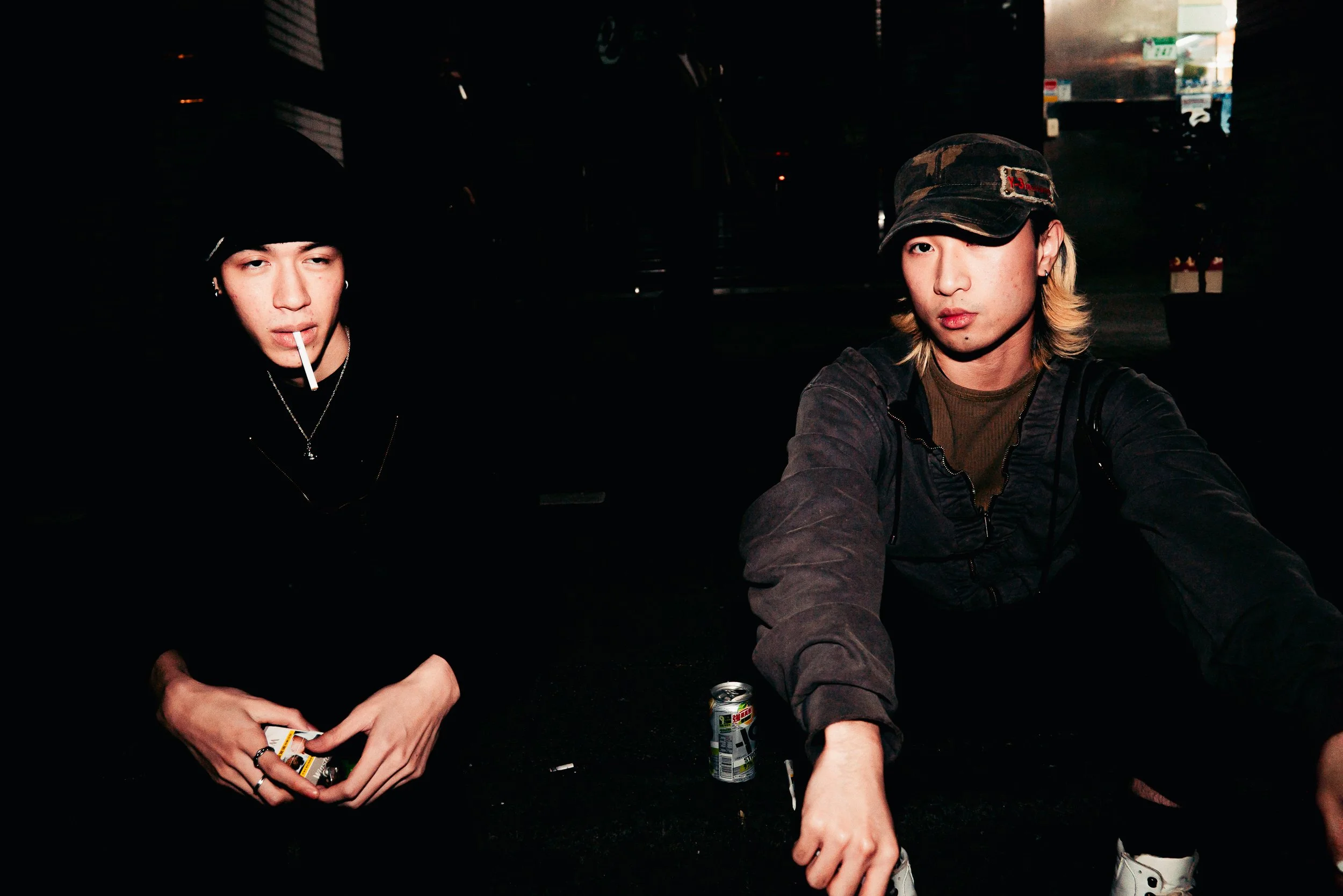 Two young men sitting in a dark outdoor setting at night. The person on the left is wearing a black beanie, earrings, and a necklace, holding playing cards and a cigarette in his mouth. The person on the right has long blond hair, wears a camouflage 
