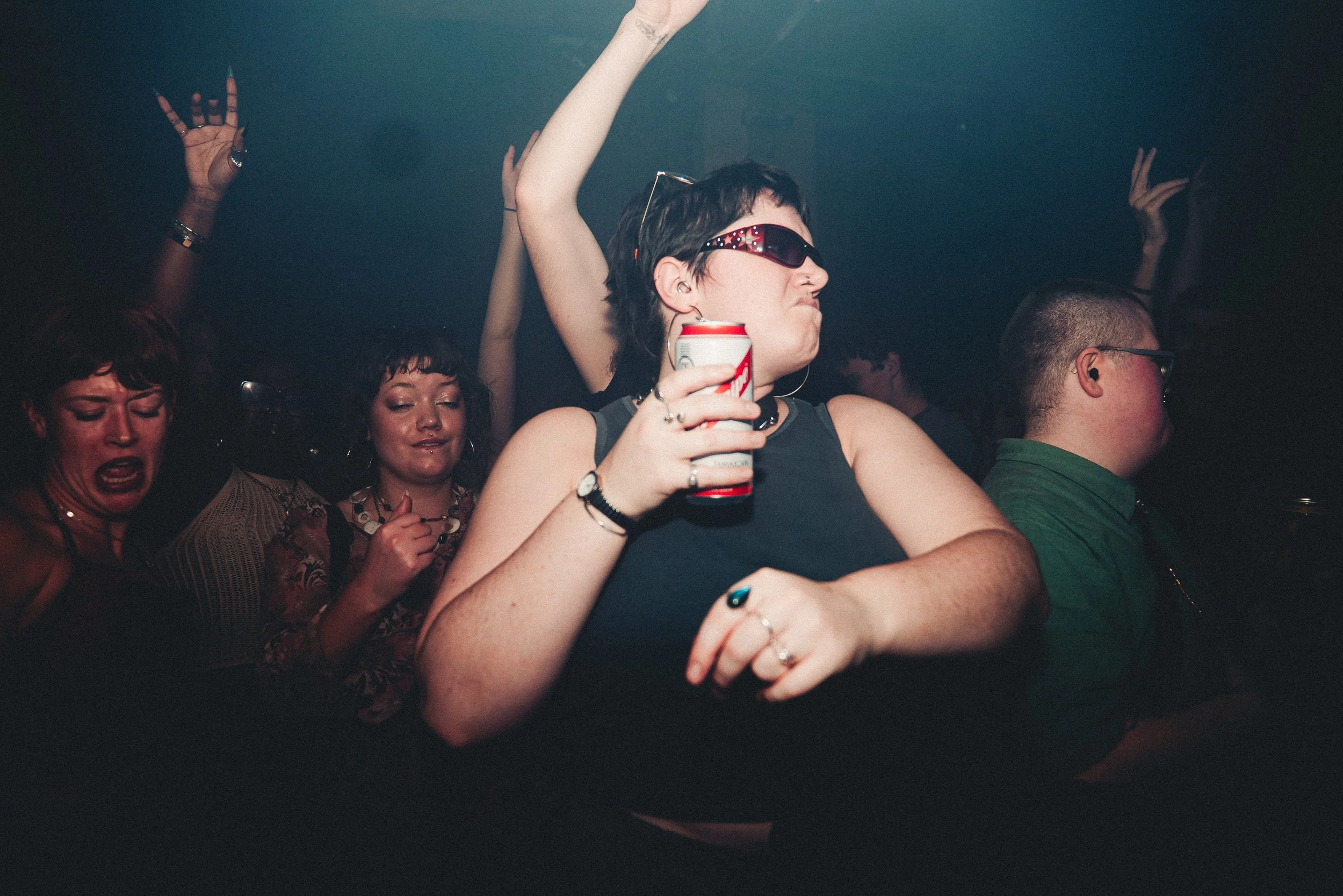 People enjoying music and dancing in a dimly lit nightclub, with some individuals raising their hands and holding drinks.