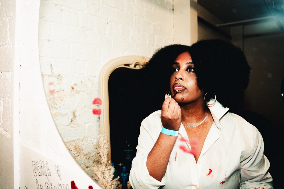 A woman with black, voluminous Afro hairstyle applying lipstick in front of a mirror.