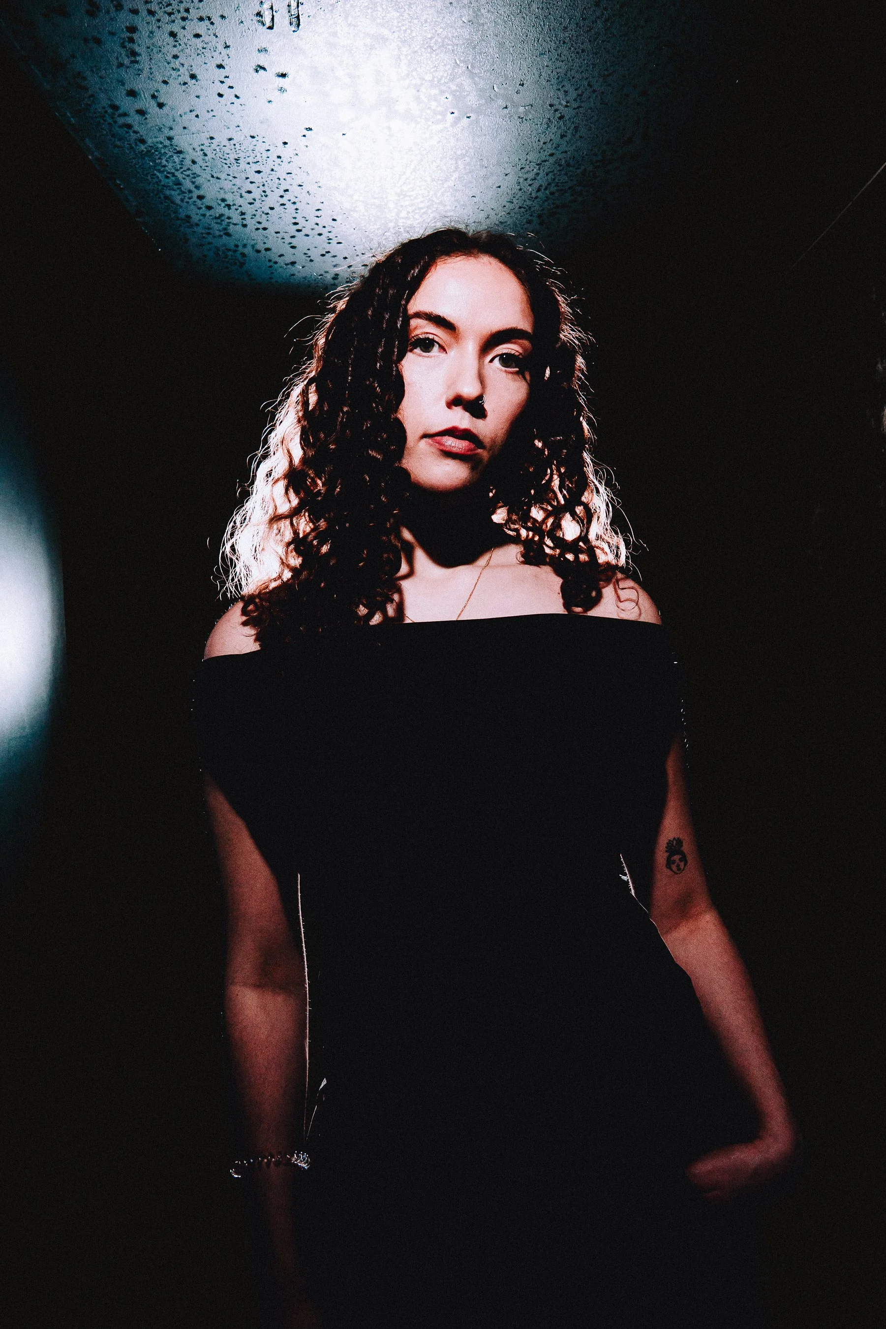 A woman with curly dark hair and fair skin poses in a black off-shoulder top against a black background with a textured ceiling reflection.