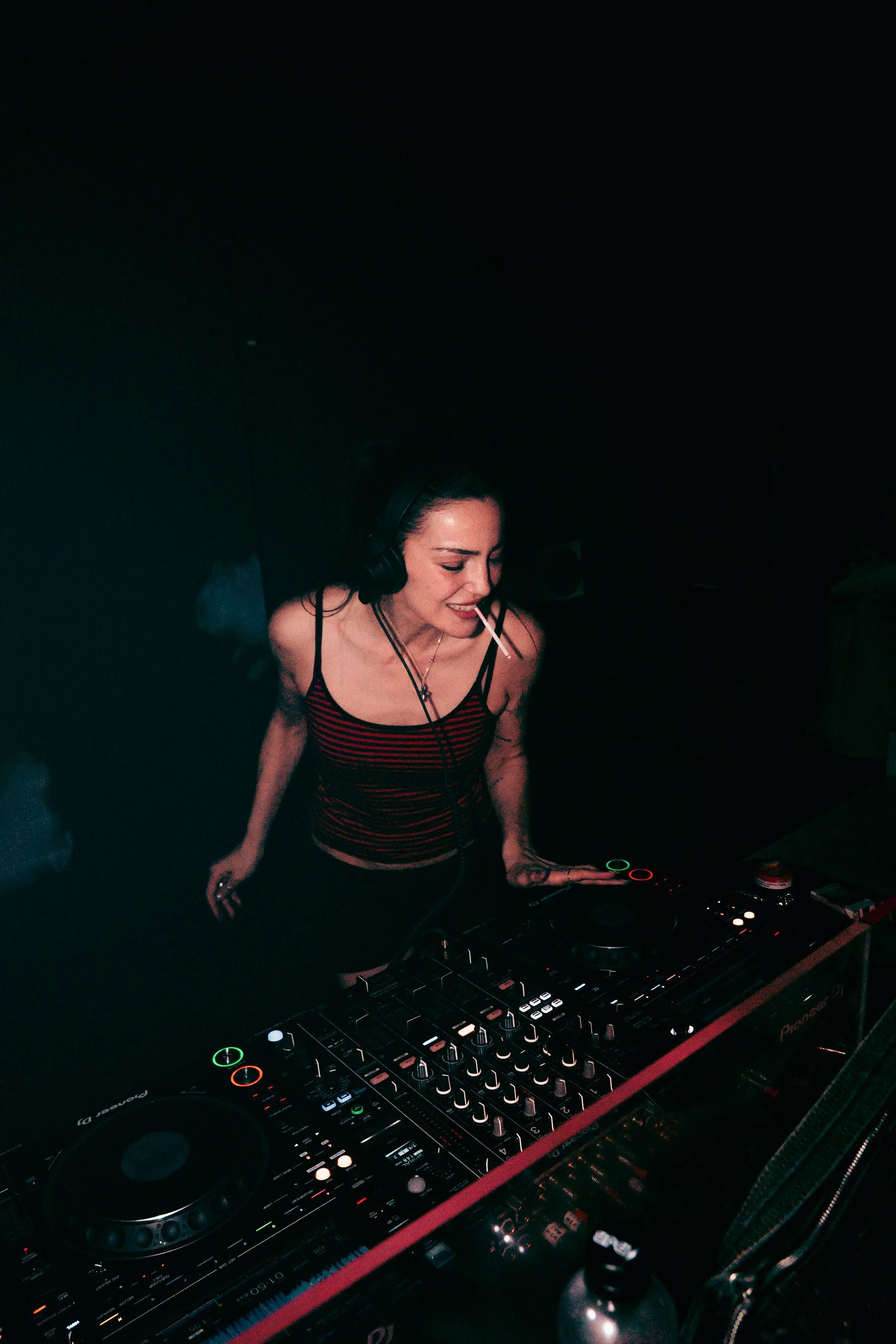A woman with black headphones and a striped tank top DJing at a club, smiling while using a Pioneer DJ controller under dim lighting.