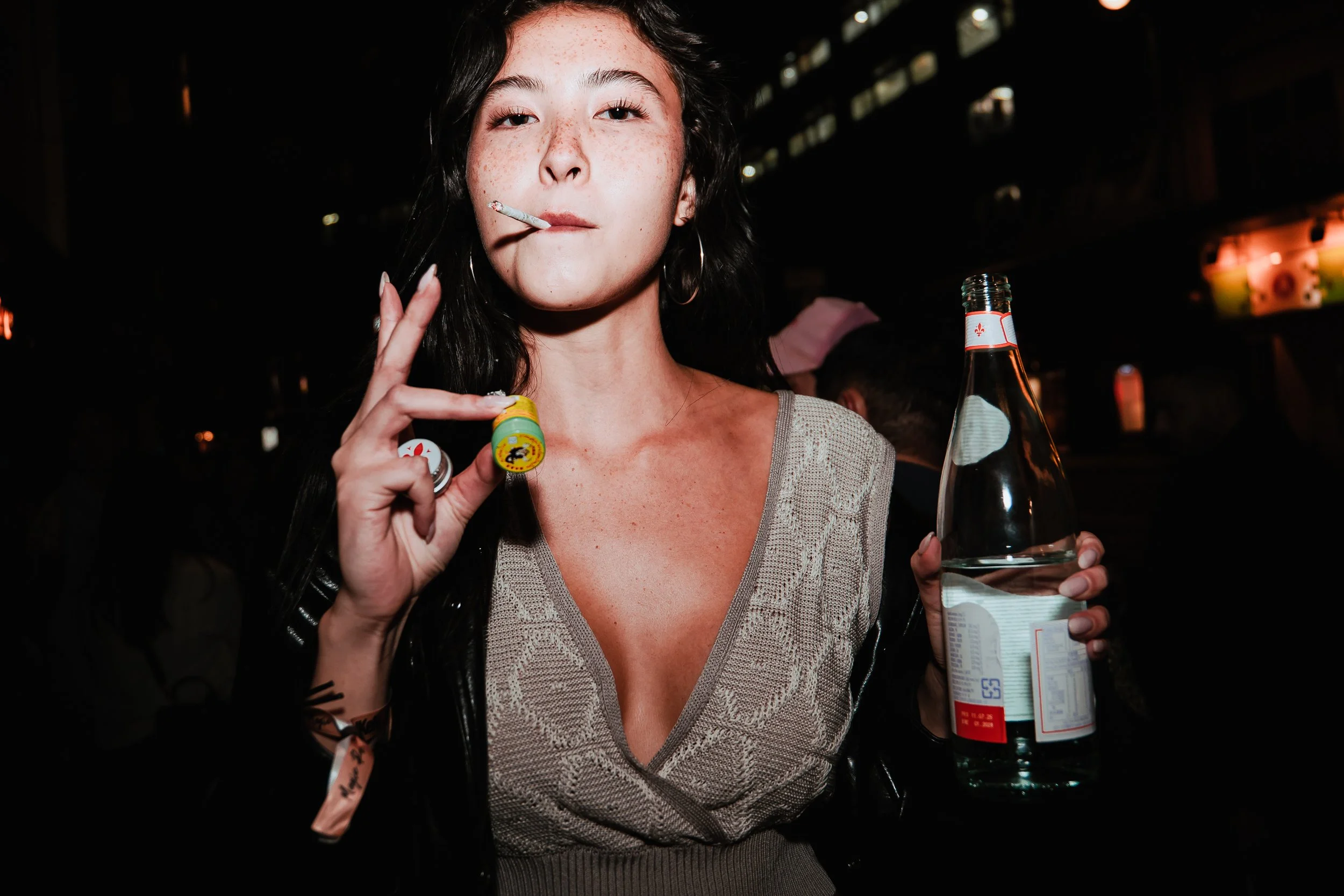 A young woman with dark hair and freckles holding a cigarette in her mouth and a glass bottle in her right hand, standing in a dimly lit setting.