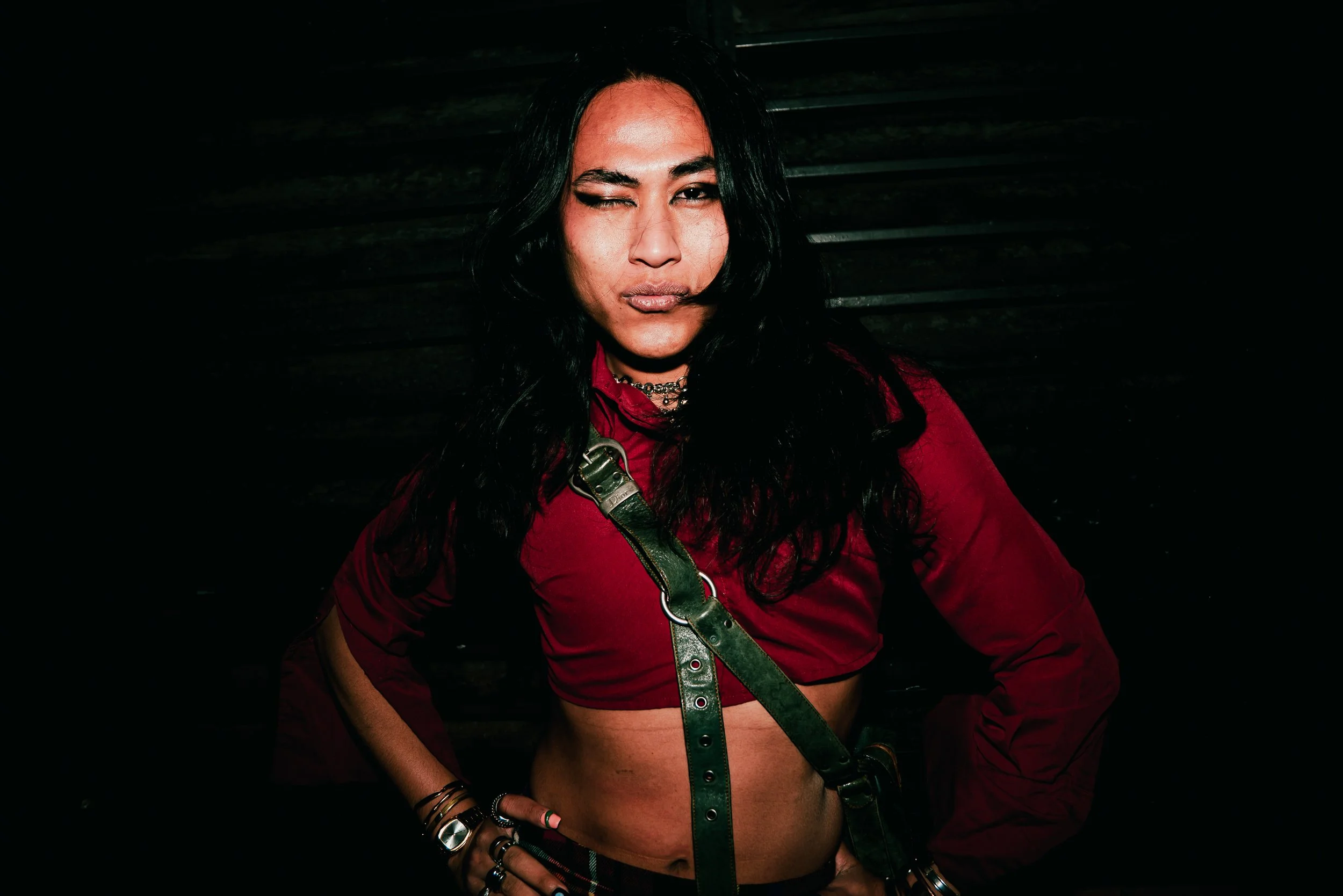 A woman with long black hair, wearing a red jacket, a green strap, and jewelry, posing with one eye closed in a dark setting.