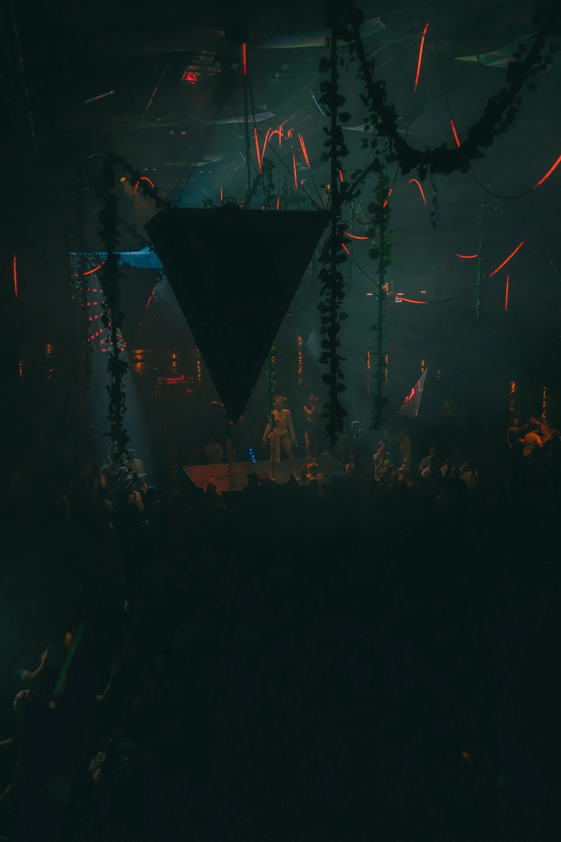 A dark indoor scene with neon lights and hanging vines, people gathered around, and what appears to be an art installation or stage setup in the center.