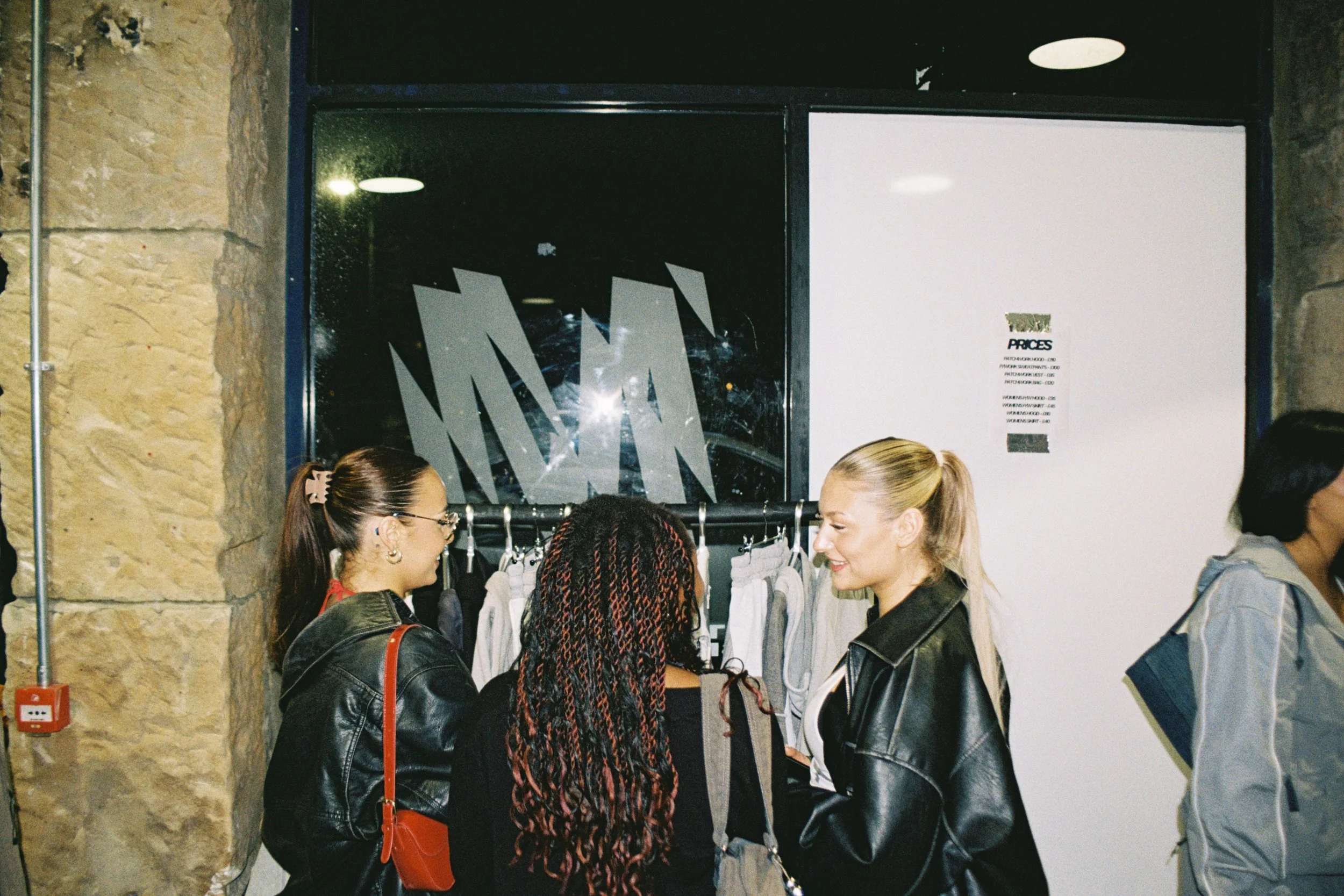 Group of people shopping and chatting indoors, with clothing on display and a price list posted on the wall.