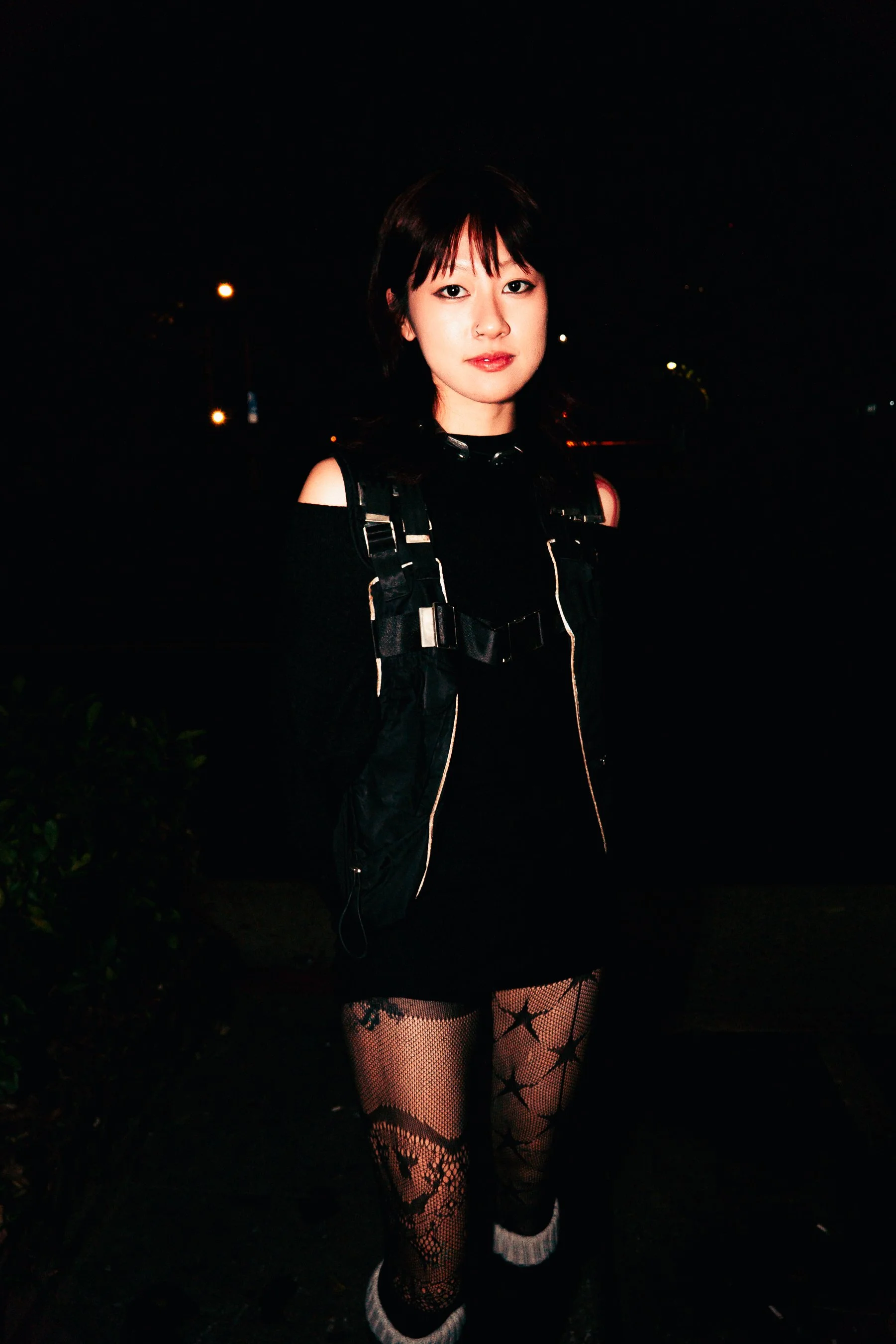 A young woman with short dark hair and makeup standing outdoors at night, wearing a black off-shoulder top, fishnet patterned stockings with stars and moon designs, and fishnet gloves with white frills at the cuffs.