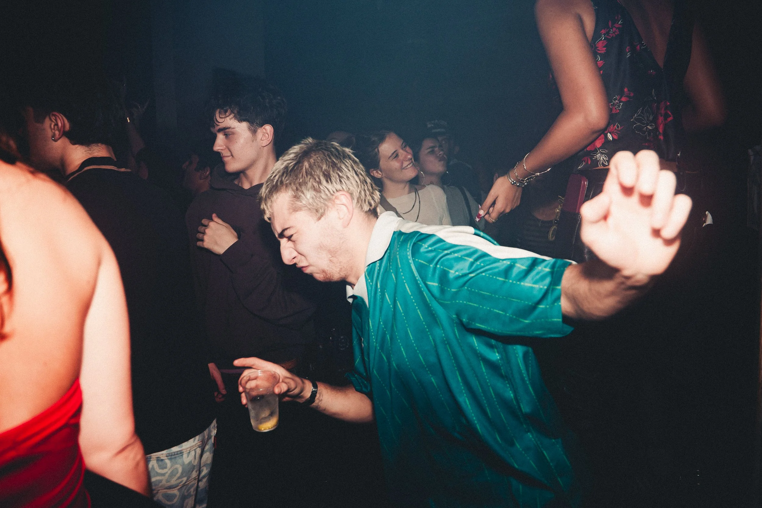 People dancing and socializing at a party in a dimly lit setting. A woman is seen holding a drink and smiling, while others are engaged in conversation or dance.