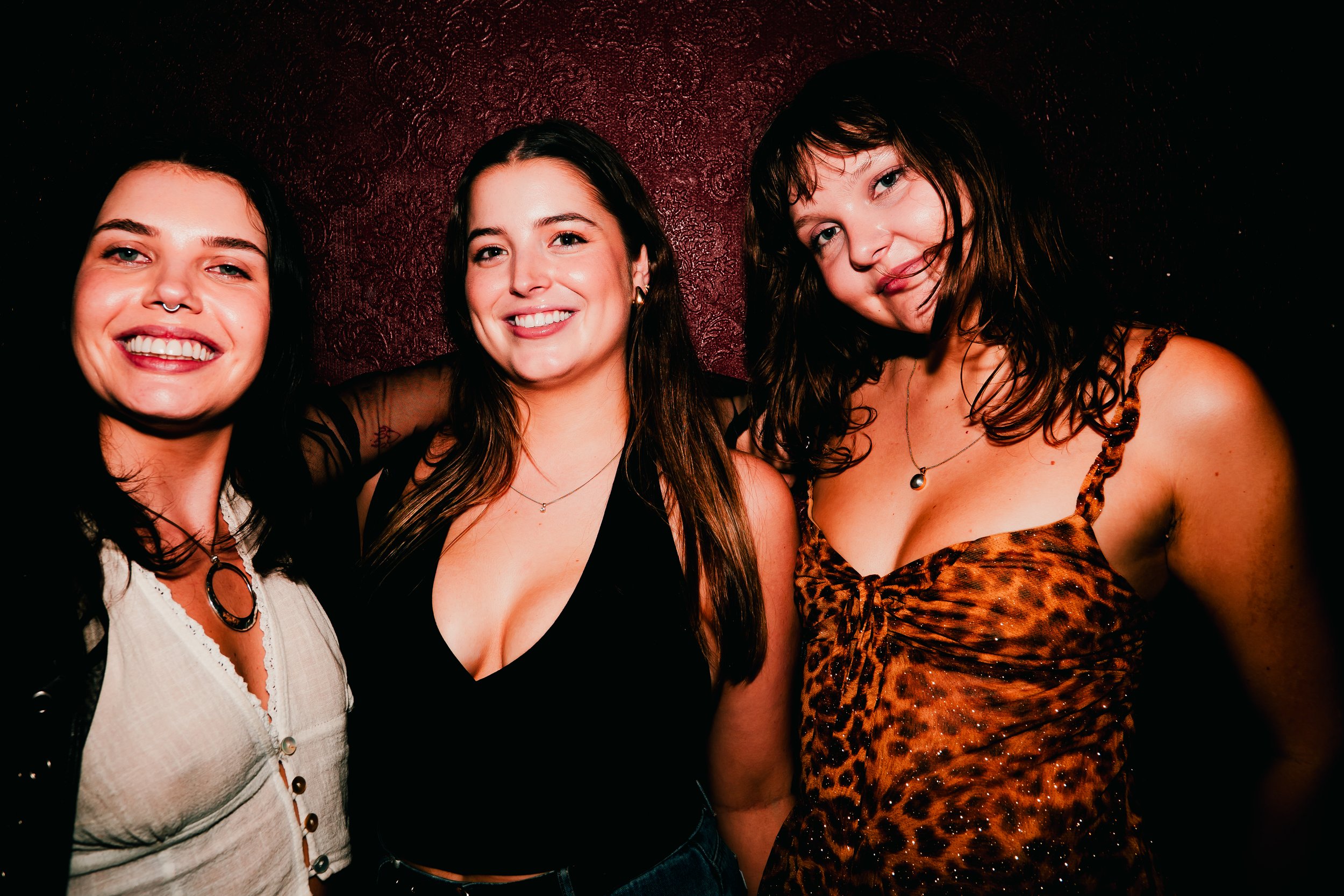 Three women smiling and posing together in a dimly lit setting with a dark textured background. The woman on the left has dark hair, a septum piercing, and is wearing a light-colored top. The woman in the middle has dark hair, is smiling widely, and 