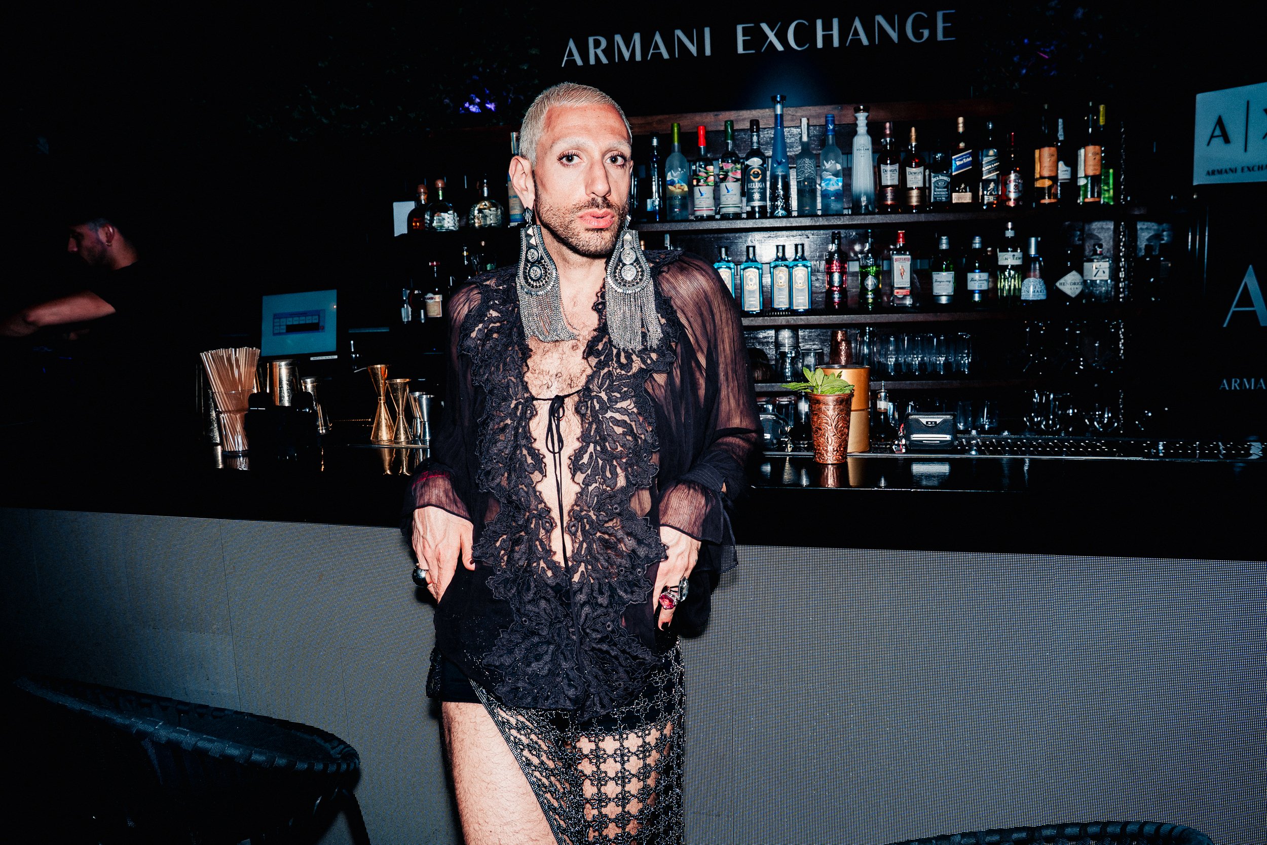 Person with short blonde hair and large earrings standing in front of a bar with bottles, wearing a black lace outfit with sheer fabric and lace designs.