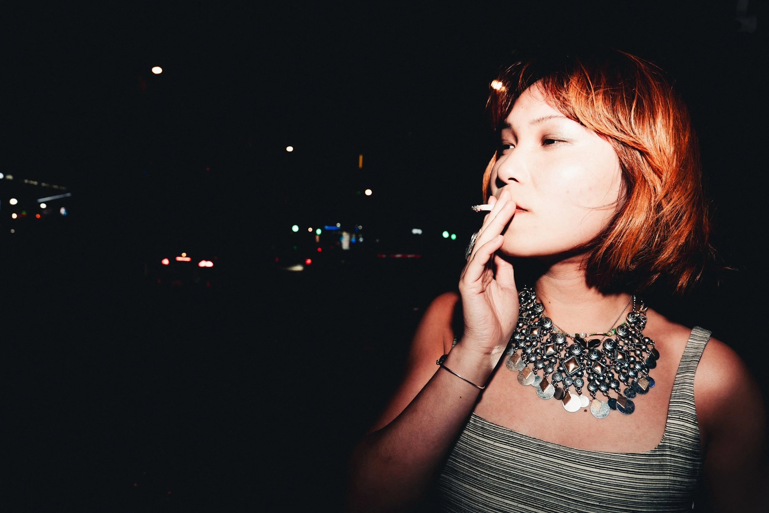 A young woman with short red hair smoking a cigarette at night with a cityscape and traffic lights in the background.